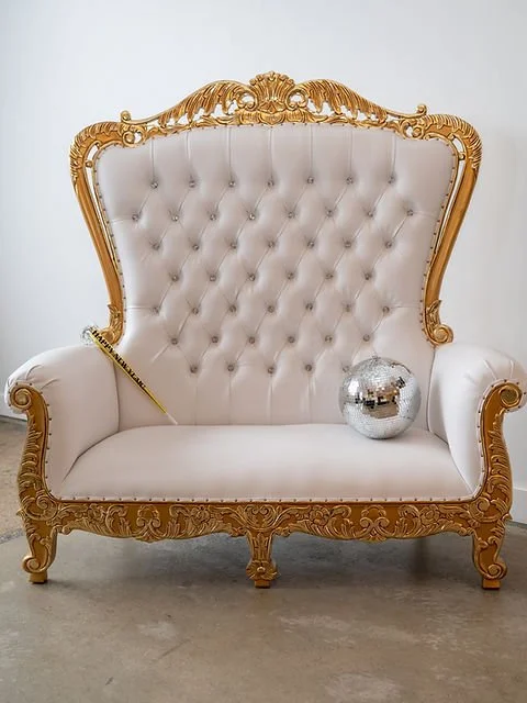 A vintage, ornate white couch with gold trim and detailing, a small disco ball on the seat, and a gold feather pen resting on the armrest.