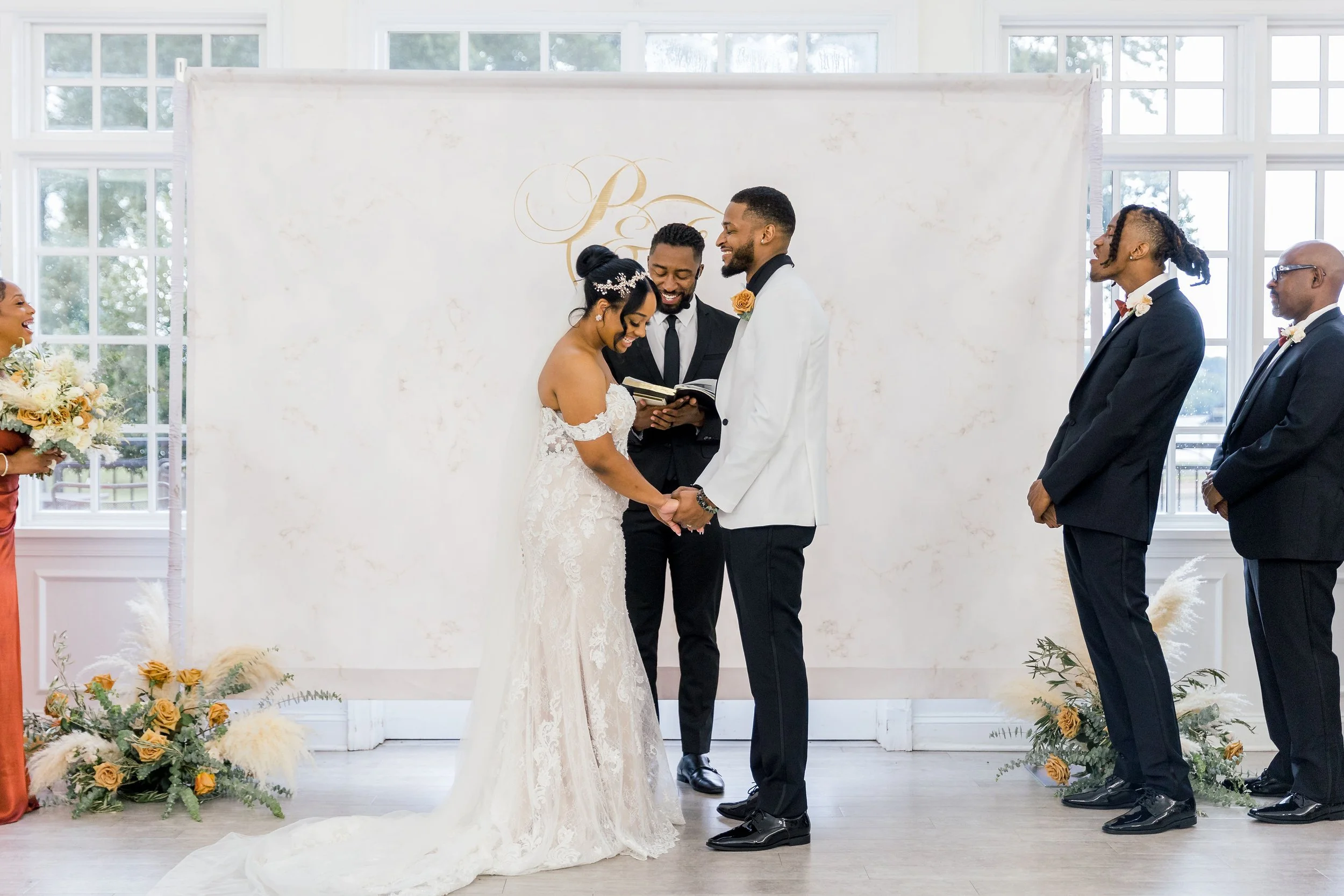 A wedding ceremony with a bride and groom holding hands and exchanging vows, surrounded by an officiant and bridesmaid and groomsmen, in a bright, indoor setting with large windows and floral decorations.