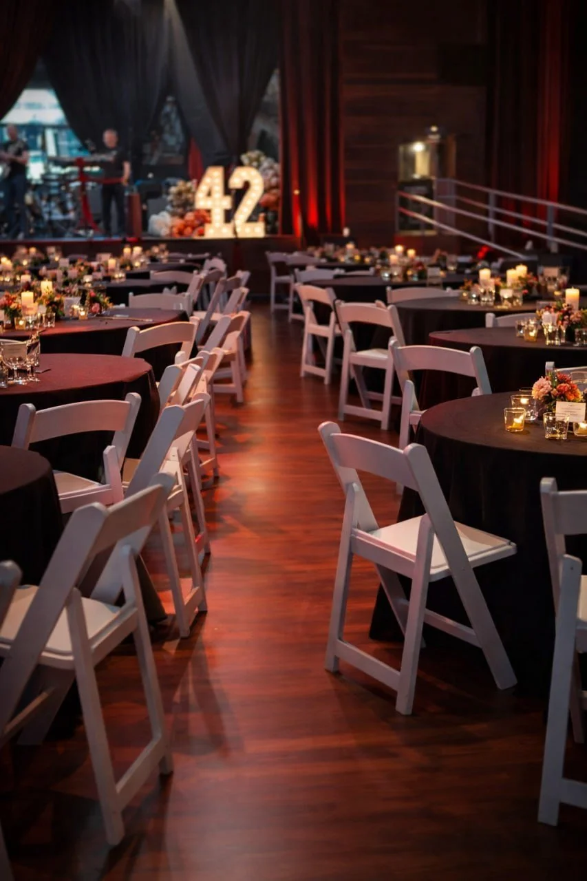 A banquet hall decorated for a celebration with multiple round tables, white chairs, floral centerpieces, lit candles, and a large illuminated '42' sign on stage in the background.