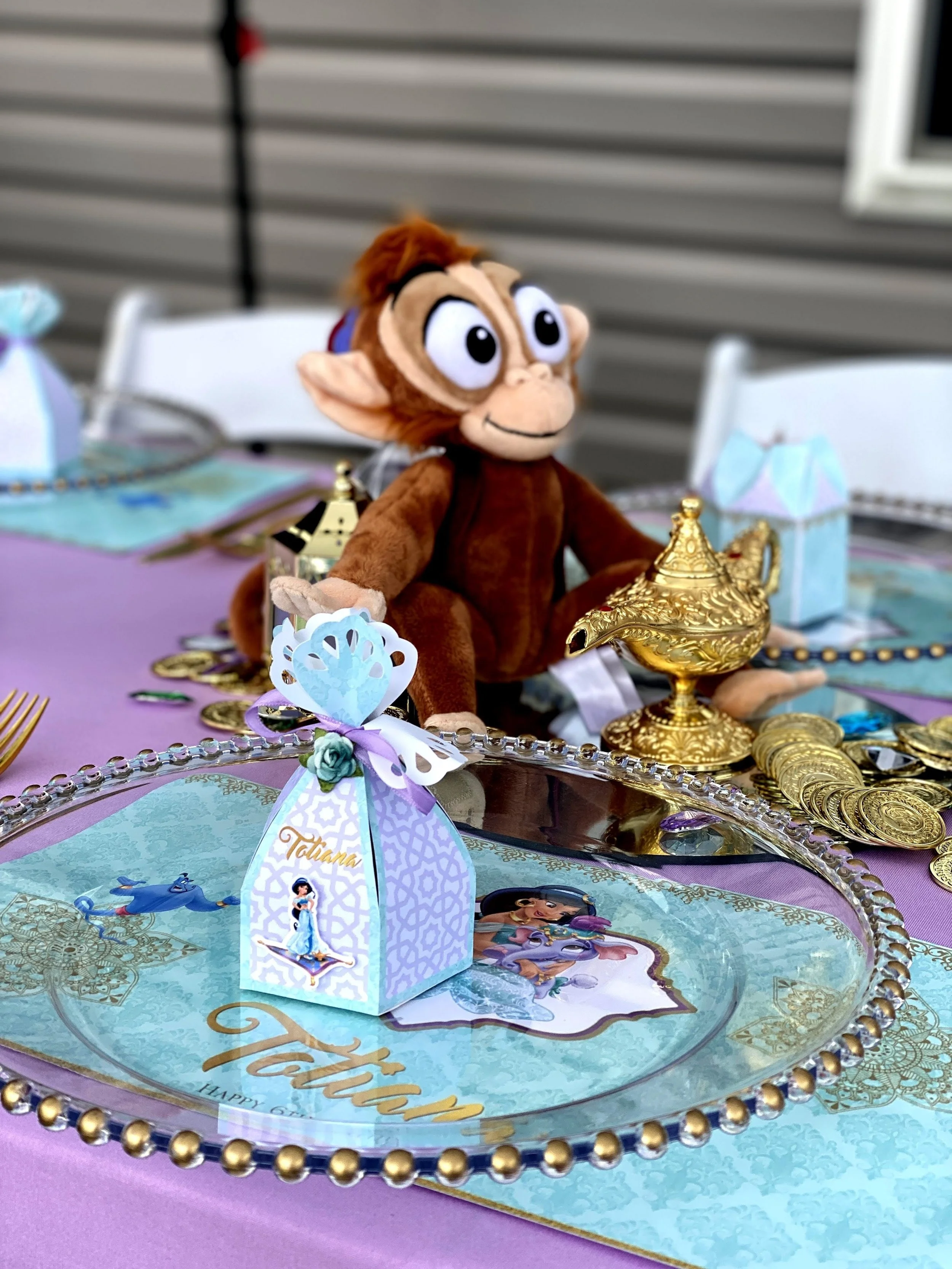 A decorated tablescape with a plush Abu from Aladdin, gold jewelry, and a themed gift box featuring Jasmine, on a pink and purple tablecloth with a 'Tatum' placement card.