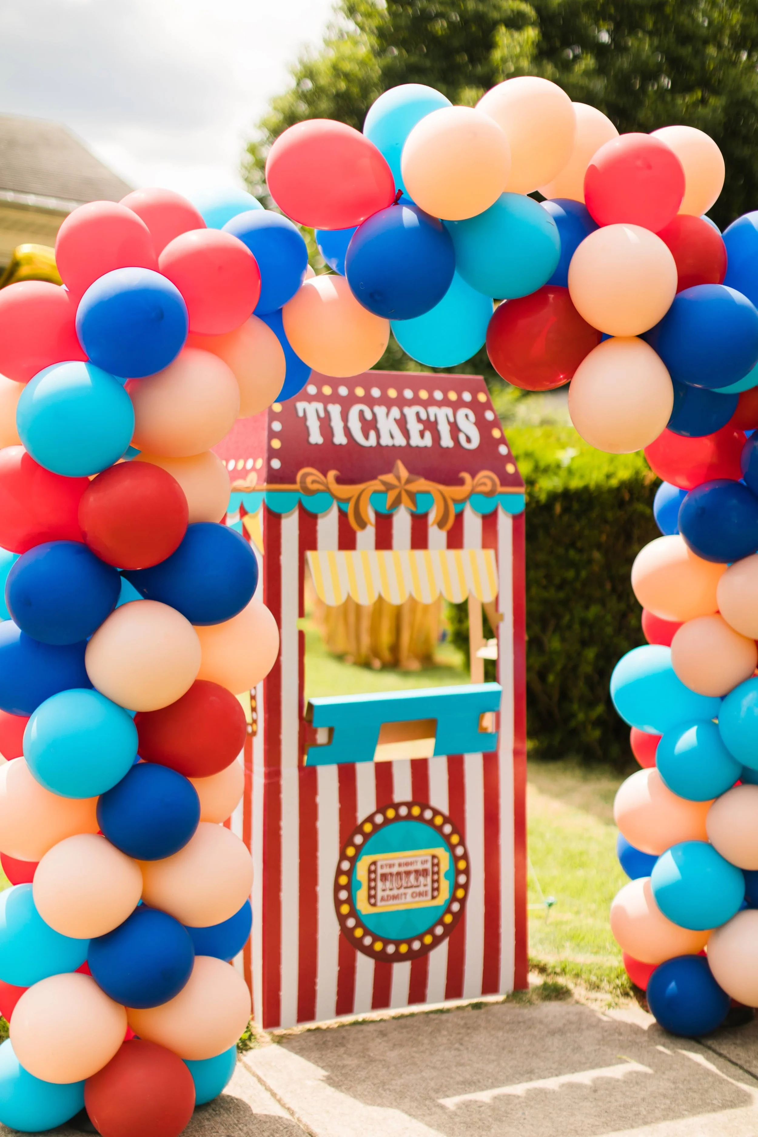 Colorful balloon arch framing a carnival ticket booth backdrop with red and white stripes, a yellow striped awning, and a sign that says 'Tickets' with circus-themed decorations.