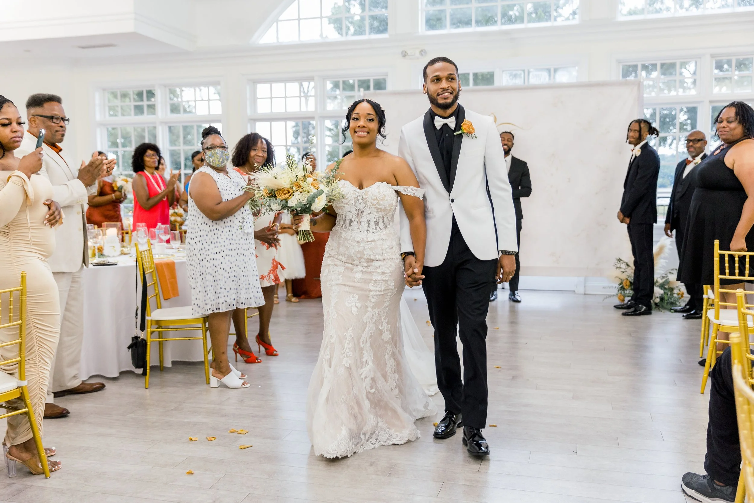 A bride and groom walking hand in hand into their wedding reception, surrounded by seated and standing guests in a bright indoor venue with large windows and gold chairs.