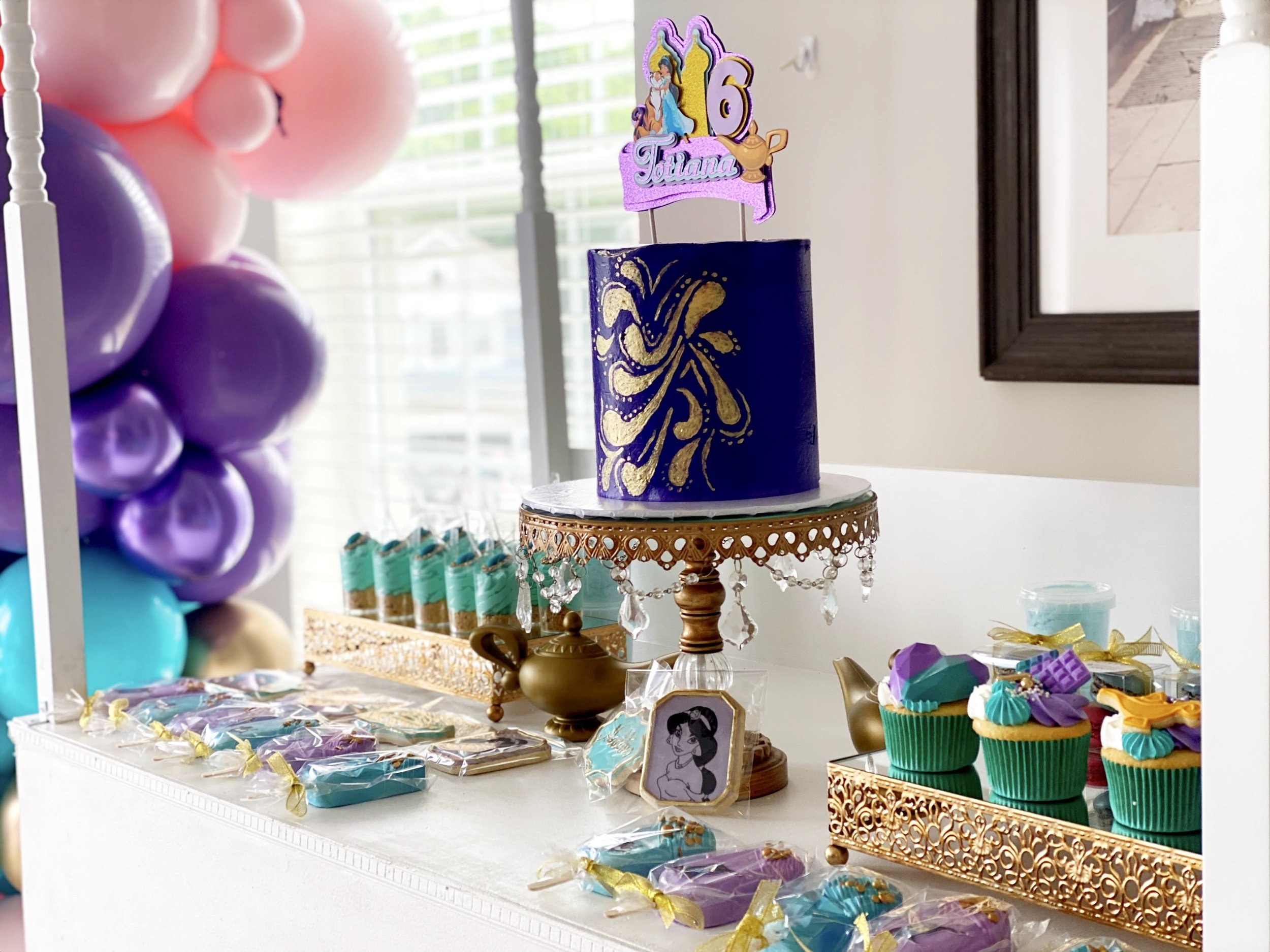 A decorated birthday table featuring a purple and gold themed cake with a Disney princess topper, surrounded by colorful cupcakes, cookies, and balloons, with a '6' candle and a sign reading 'Tiana'.