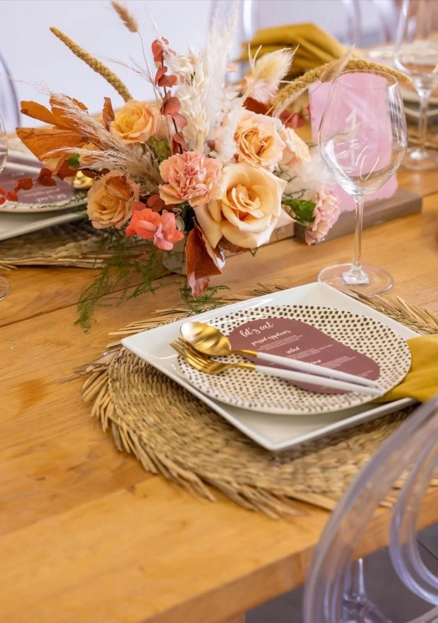 A table setting featuring a square white plate with a black dotted pattern, a gold fork and spoon, a place card, a wine glass, and a floral centerpiece with peach, pink, and cream roses, dried grasses, and green foliage.