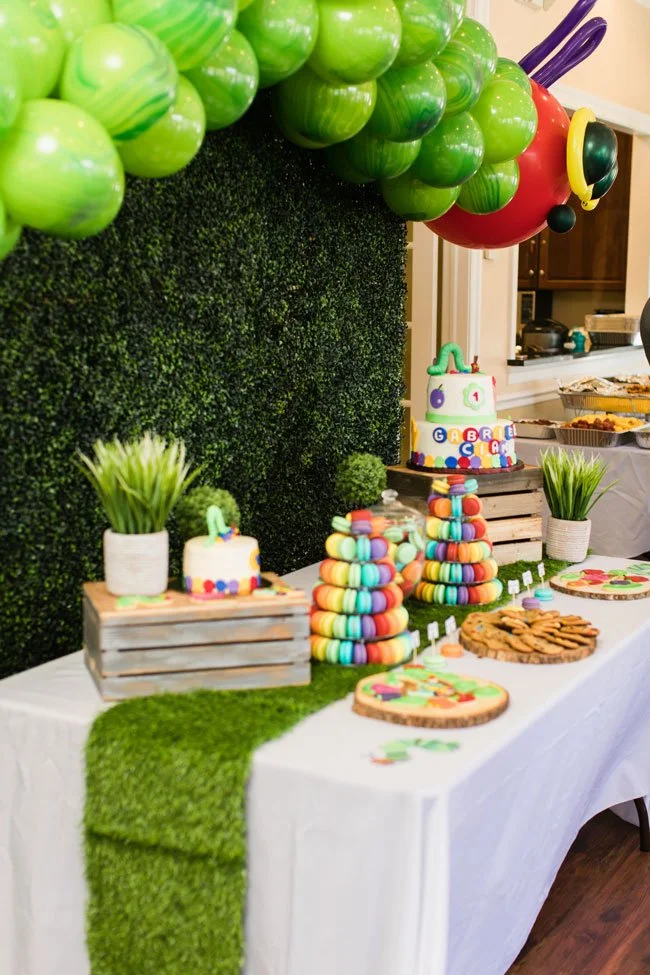 Colorful party table with green balloon decorations, potted plant centerpieces, a cake with alphabet decorations, stacks of colorful macarons, cookie treats, and themed paper cutouts, set against a green textured backdrop.