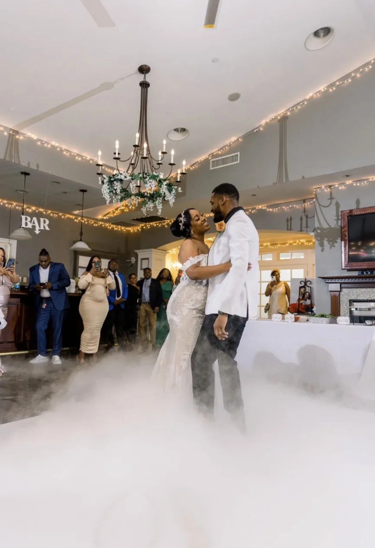 A bride and groom are dancing closely on a foggy dance floor during their wedding reception, surrounded by guests taking photos and watching.