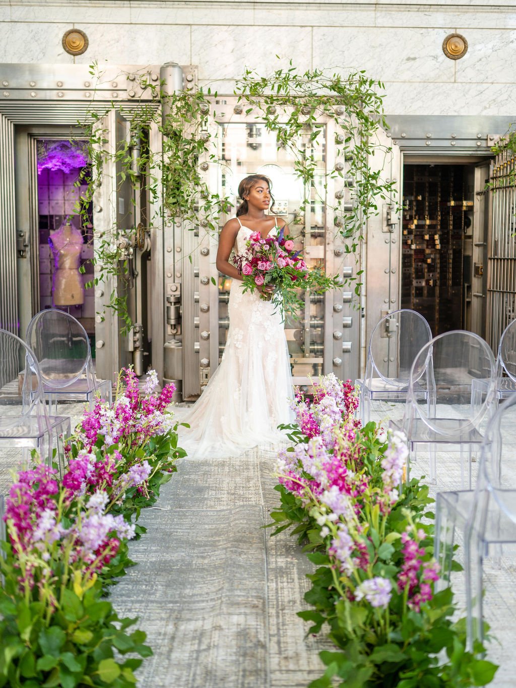 A bride in a white wedding gown holding a large bouquet of pink and purple flowers, standing amid floral arrangements with purple, pink, and white flowers, in a modern setting with metallic walls and transparent chairs.