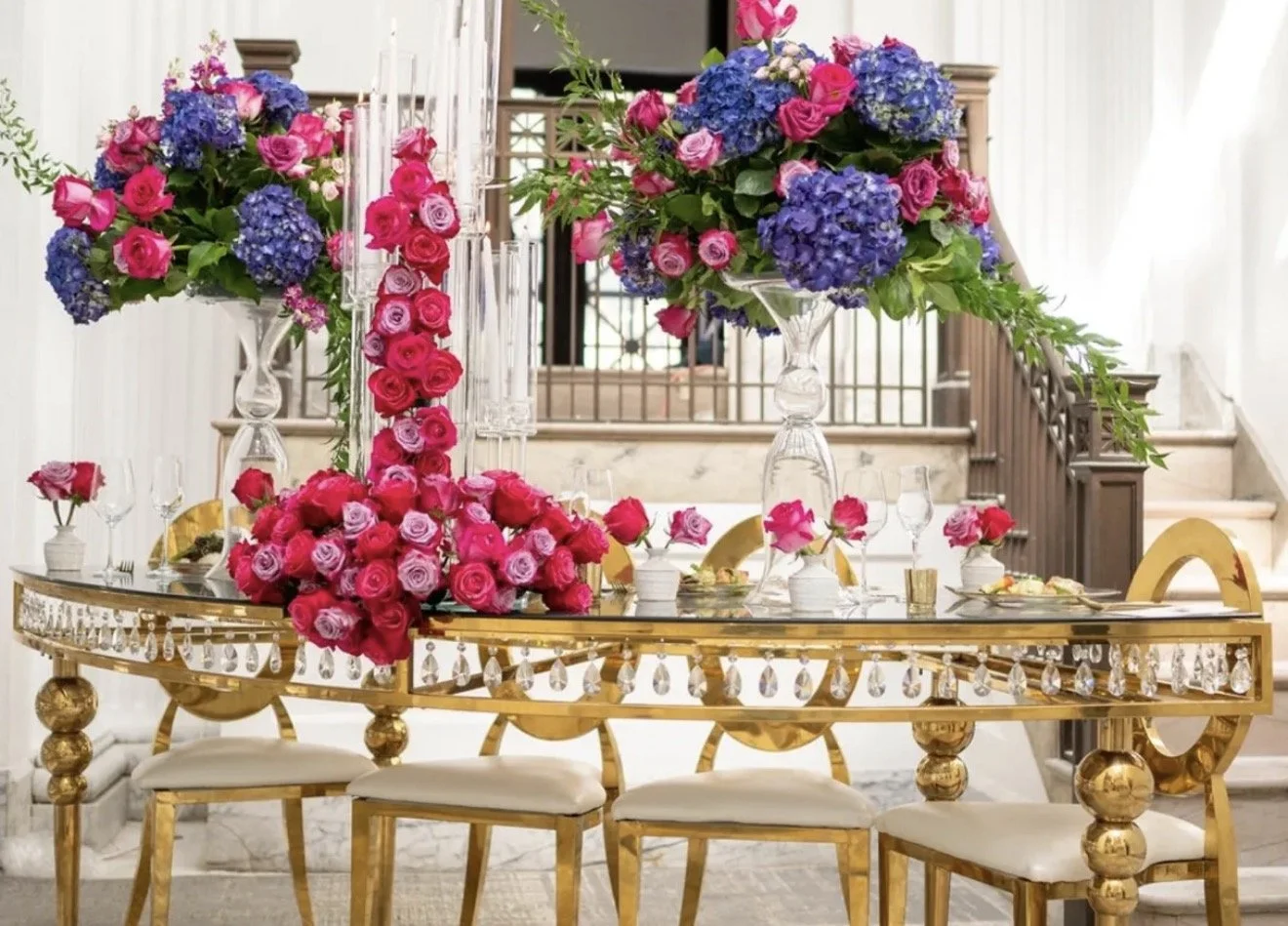 Elegant dining table with gold accents, decorated with vibrant pink and purple flower arrangements, glassware, and candles in a luxurious setting with staircase in the background.