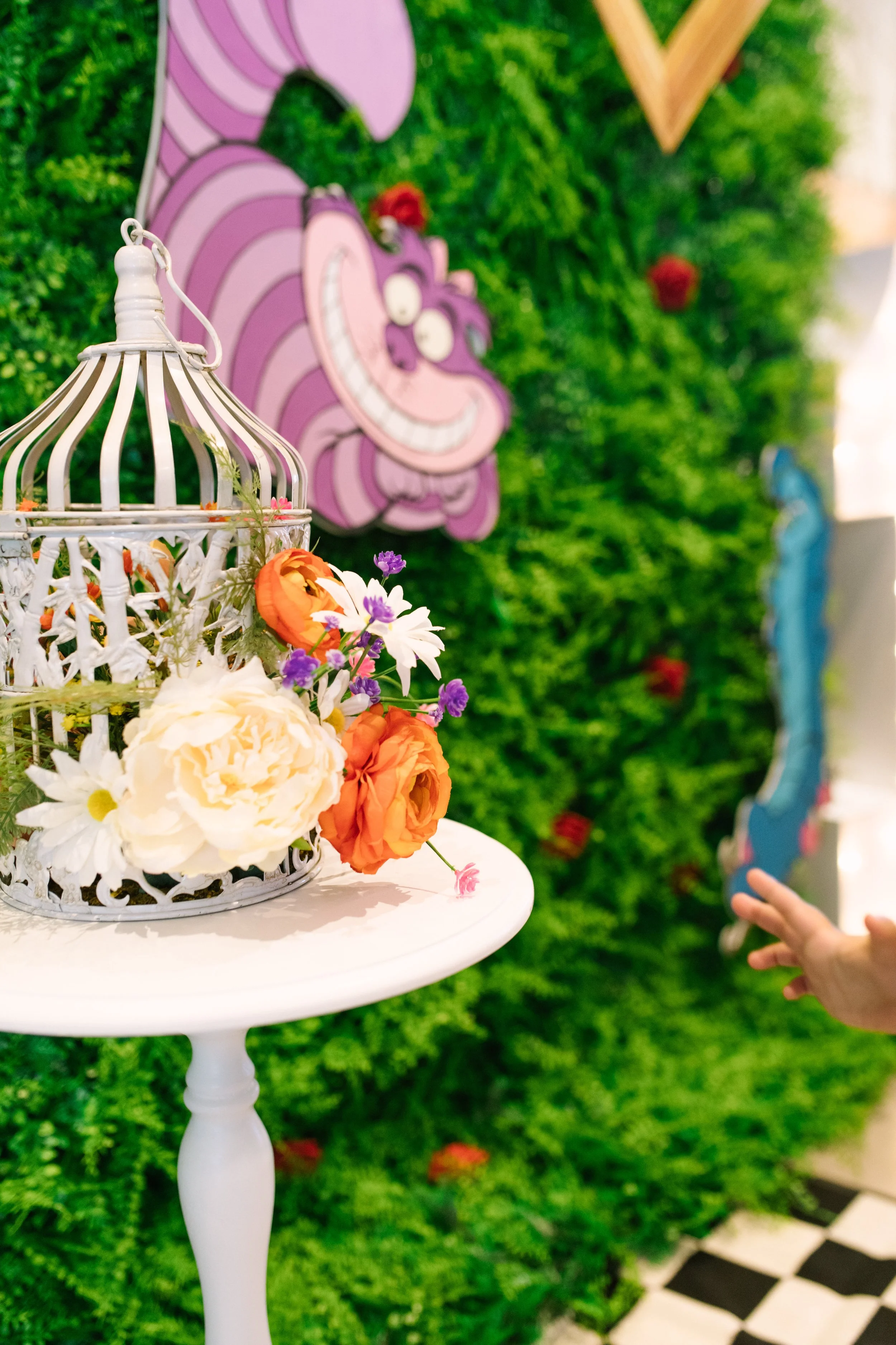 A decorative display featuring a white birdcage with artificial flowers on a white side table, with a colorful backdrop of greenery and cartoon characters.