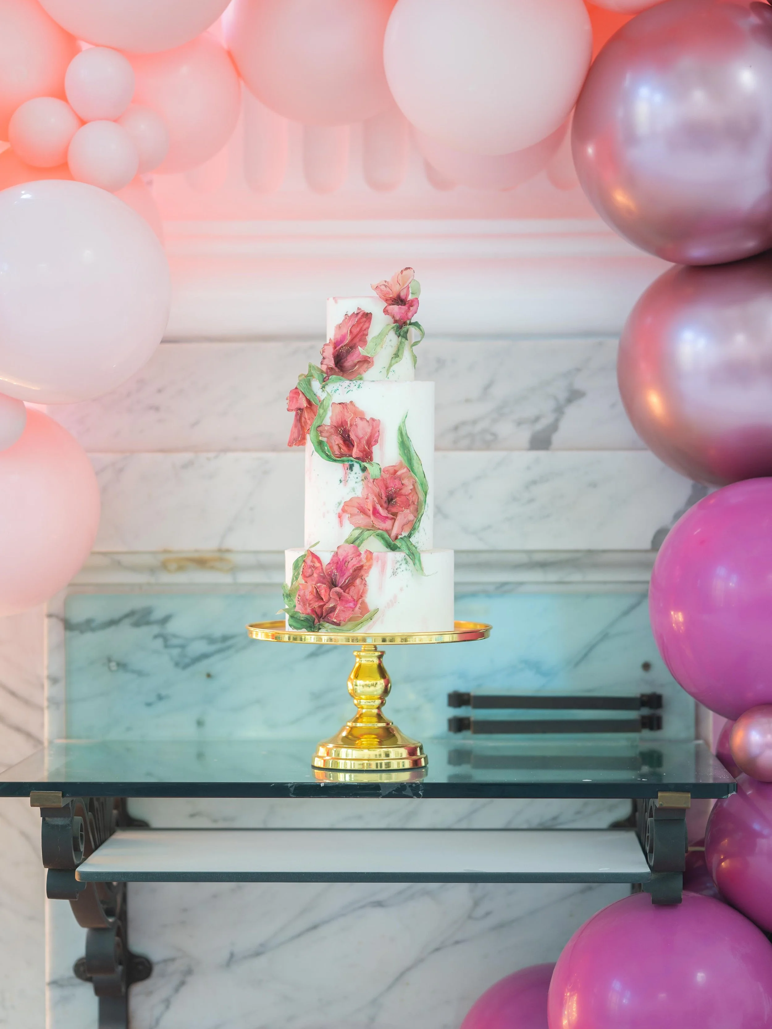 A white, multi-tier cake with pink flower decorations on a gold cake stand, surrounded by pink and purple balloons on a marble and blue background.