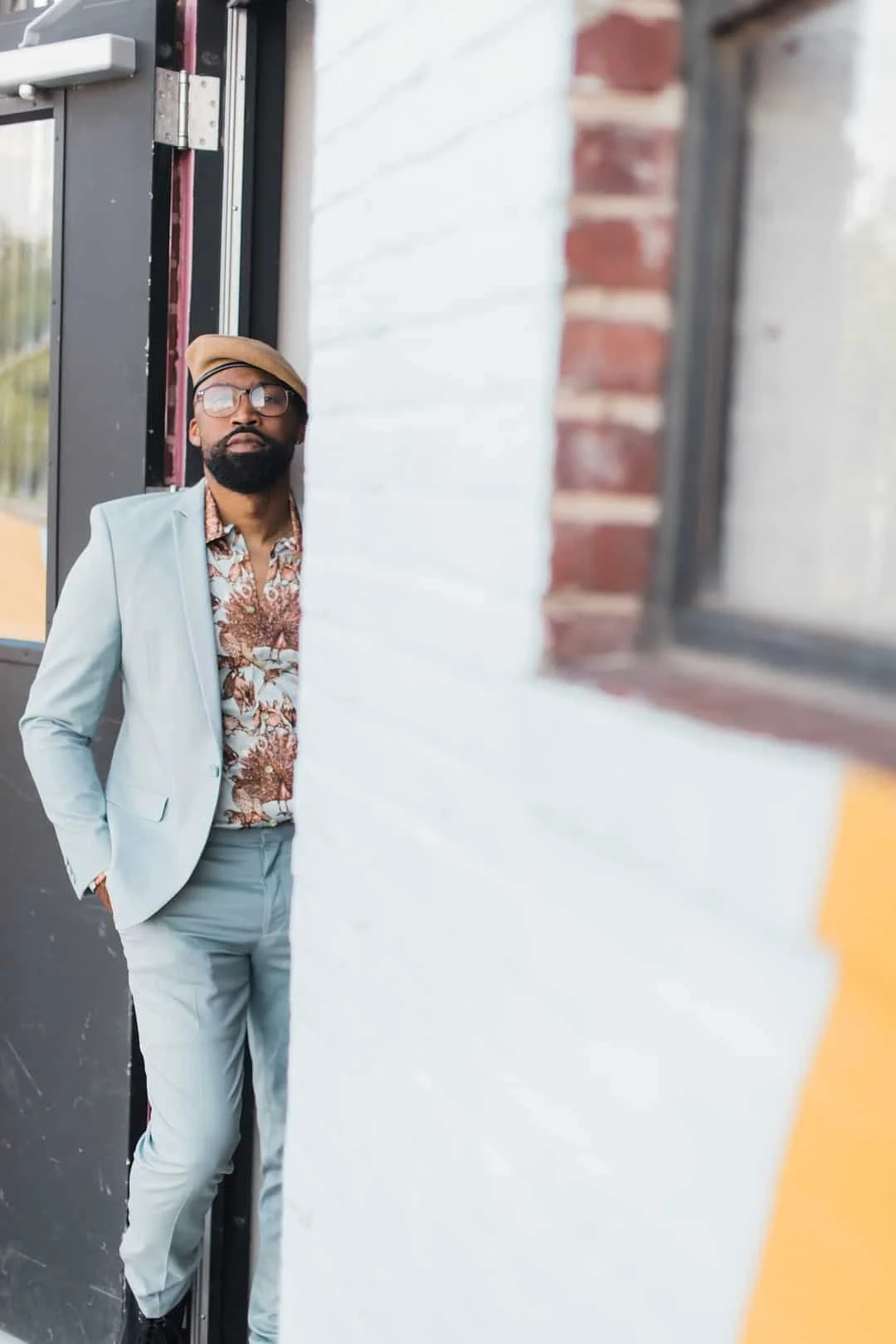 A man in a light blue suit, patterned shirt, beige beret, and glasses leans against a wall near a doorway, with a brick wall and window in the background.
