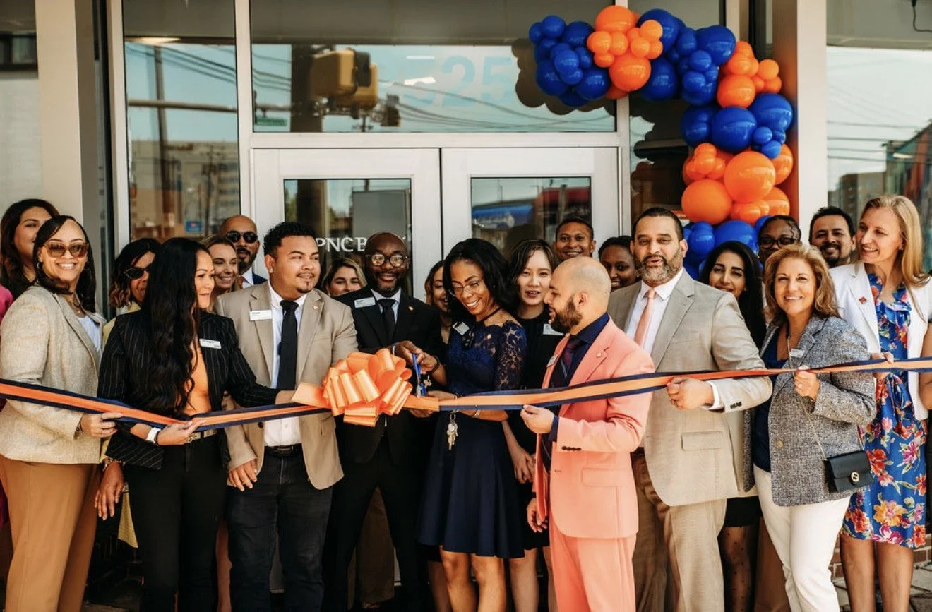 A group of diverse people at a ribbon-cutting ceremony outside a new business storefront decorated with orange and blue balloons, celebrating the grand opening.