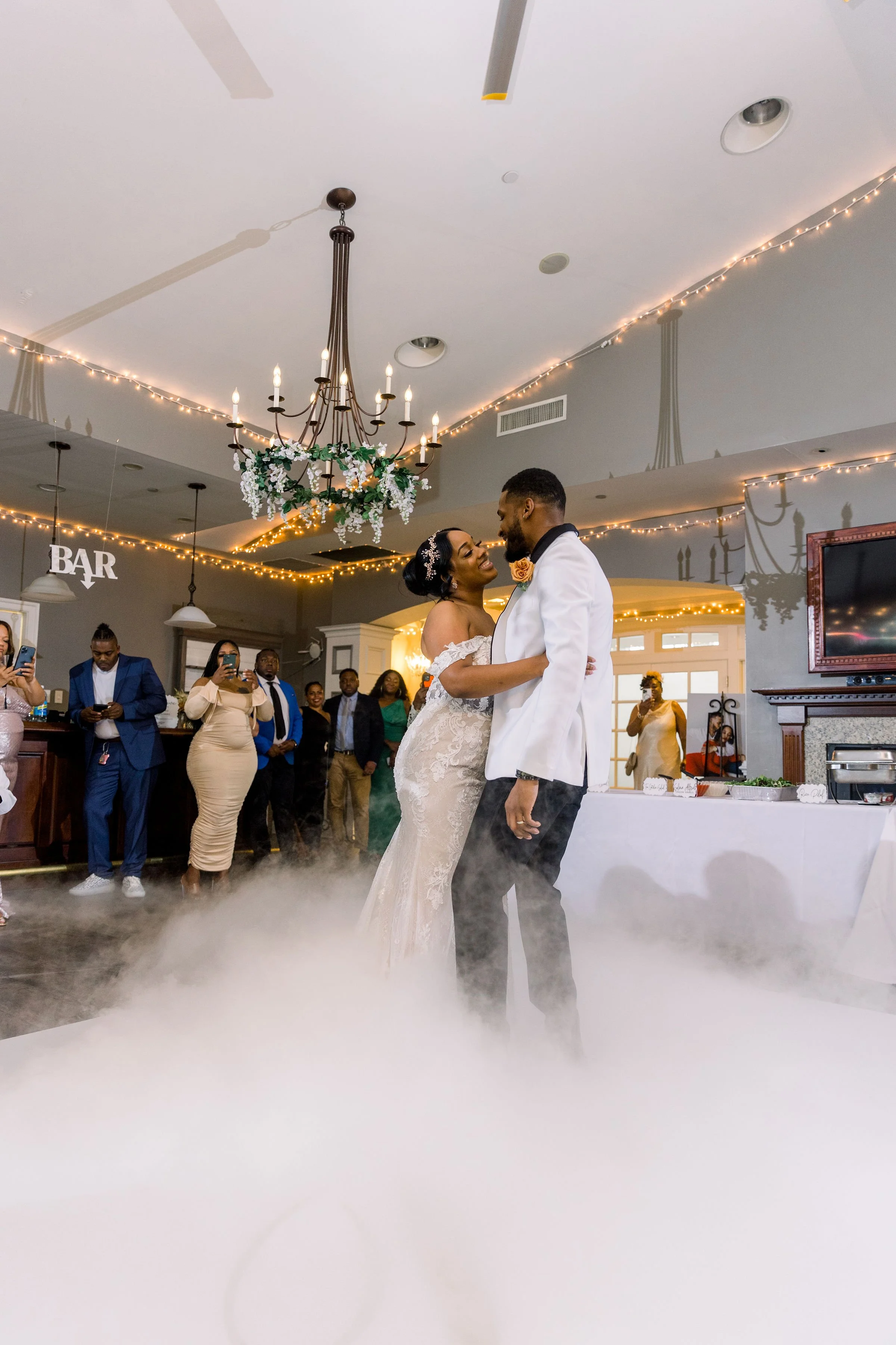 Couple dancing at their wedding reception surrounded by family and friends, with a fog effect on the dance floor, decorated with string lights and a chandelier overhead.