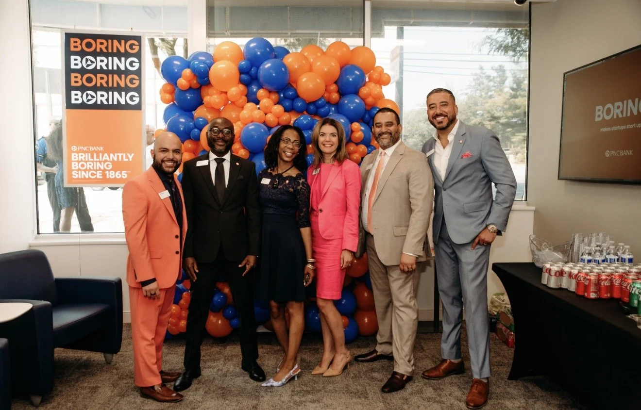 Six professionally dressed people posing in front of a colorful balloon backdrop at a corporate event, with a banner on the side reading 'Boring Boring Boring' and a table of beverages on the right.