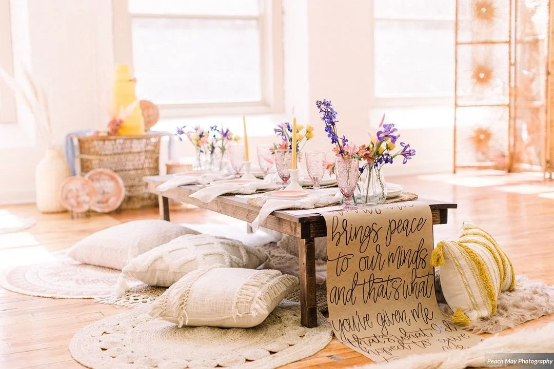 A cozy indoor setting with a low wooden table decorated with glassware, flowers, and candles, surrounded by cushions and throw pillows on the floor, with a soft pink and natural color scheme.