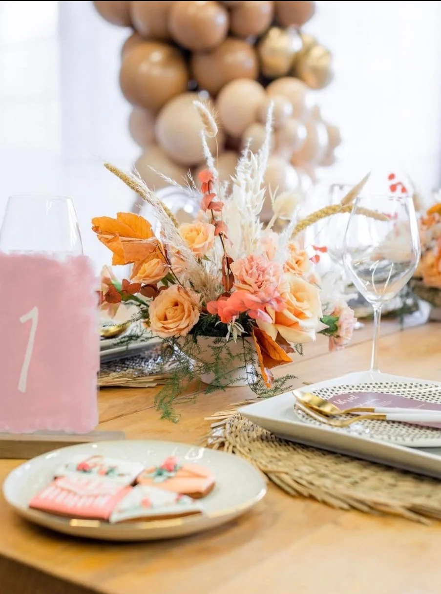 A decorated table with a pink number one candle, floral centerpiece with peaches, pink, orange, and white flowers, set with gold utensils, glassware, plates, and festive decorations.