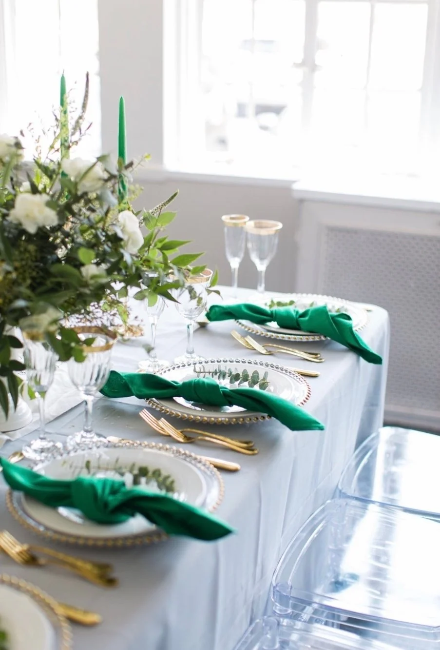 Elegant dining table set with white tablecloth, gold utensils, clear glassware with gold rims, green napkins tied around white plates with pearl-beaded edges, and a floral centerpiece with white flowers and green leaves, near large windows letting in