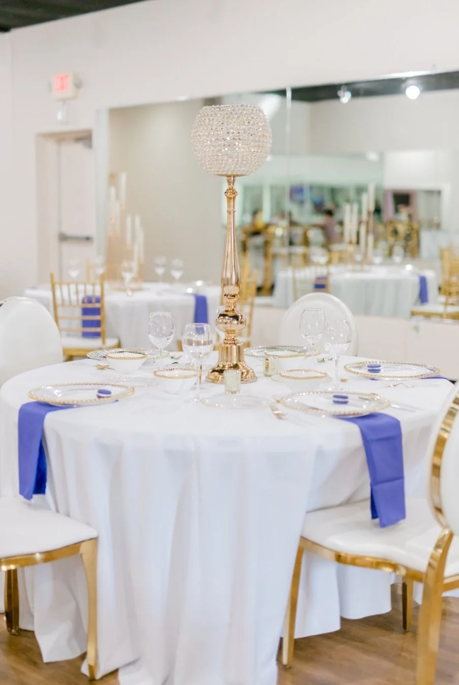 Elegant banquet table with white tablecloth, gold accents, and a tall crystal chandelier centerpiece, set in a spacious event hall.