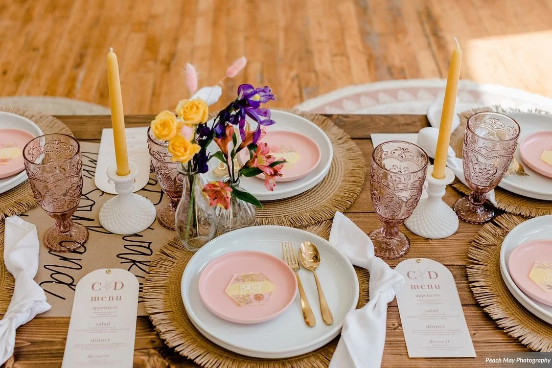 A round wooden table set for a meal with pink and white plates, gold utensils, pink glassware, and decorative candles, with a floral centerpiece.