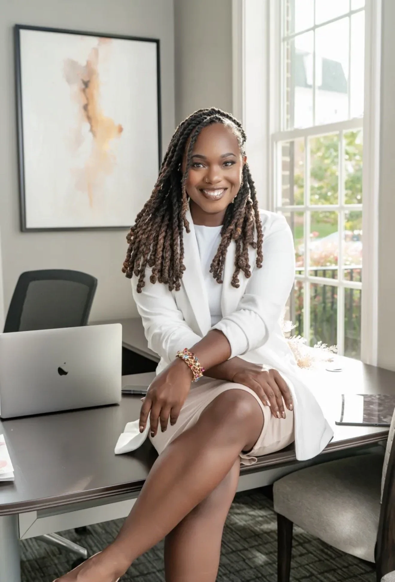 A woman with long twists in her hair, smiling, sitting on a desk in an office, wearing a white blazer and beige skirt, with a laptop and colorful bracelet, with a large window and abstract art on the wall behind her.