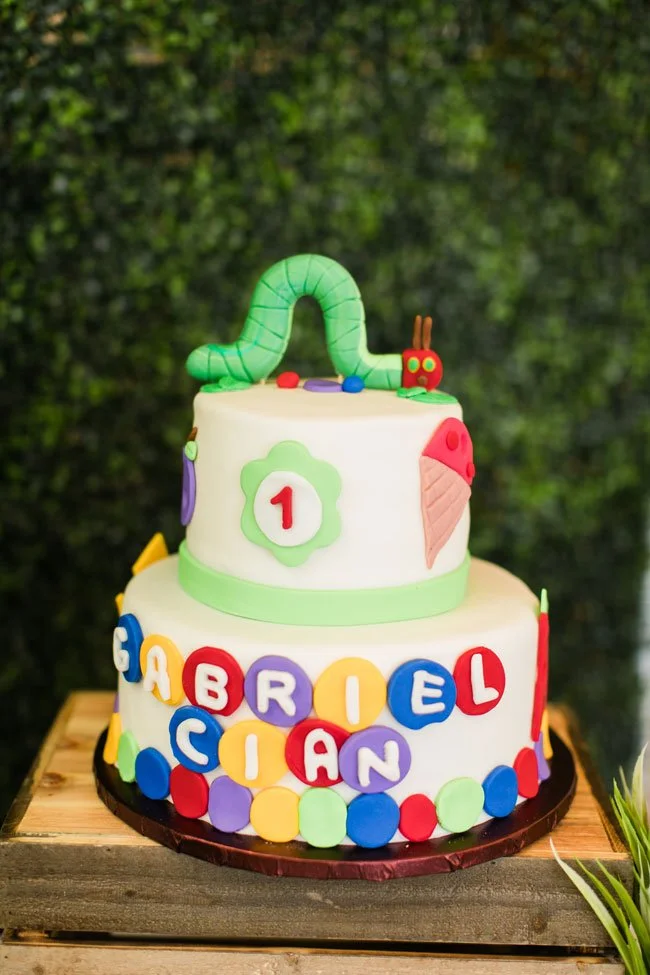 Colorful two-tiered cake decorated with a green worm, a ladybug, an ice cream cone, and multi-colored letters spelling 'Gabriel Ciean' on the front, at a birthday celebration.