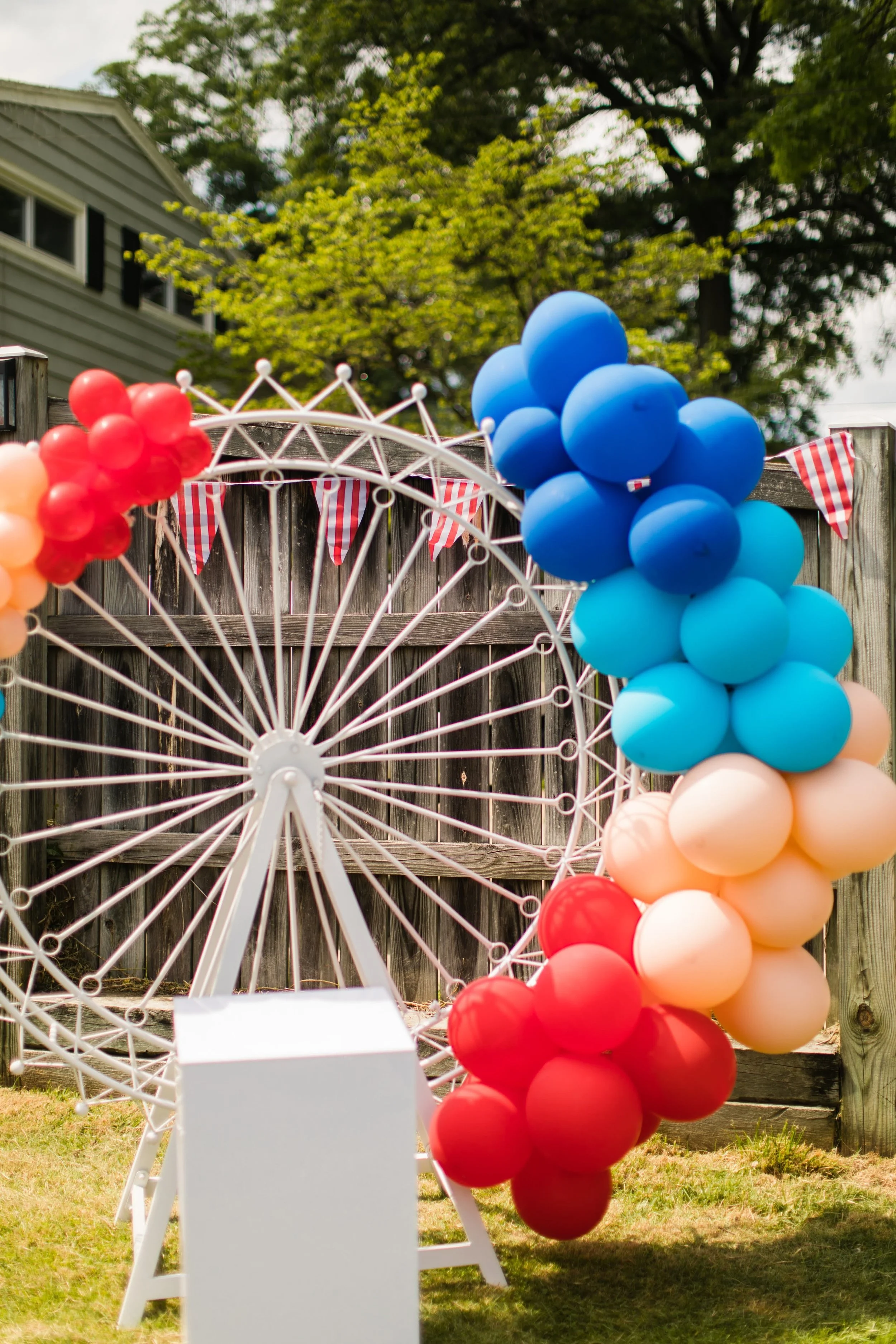 Colorful balloons arranged in red, blue, peach, and teal around a white Ferris wheel structure with red, white, and striped bunting hanging above, set outdoors with a backyard fence and trees in the background.