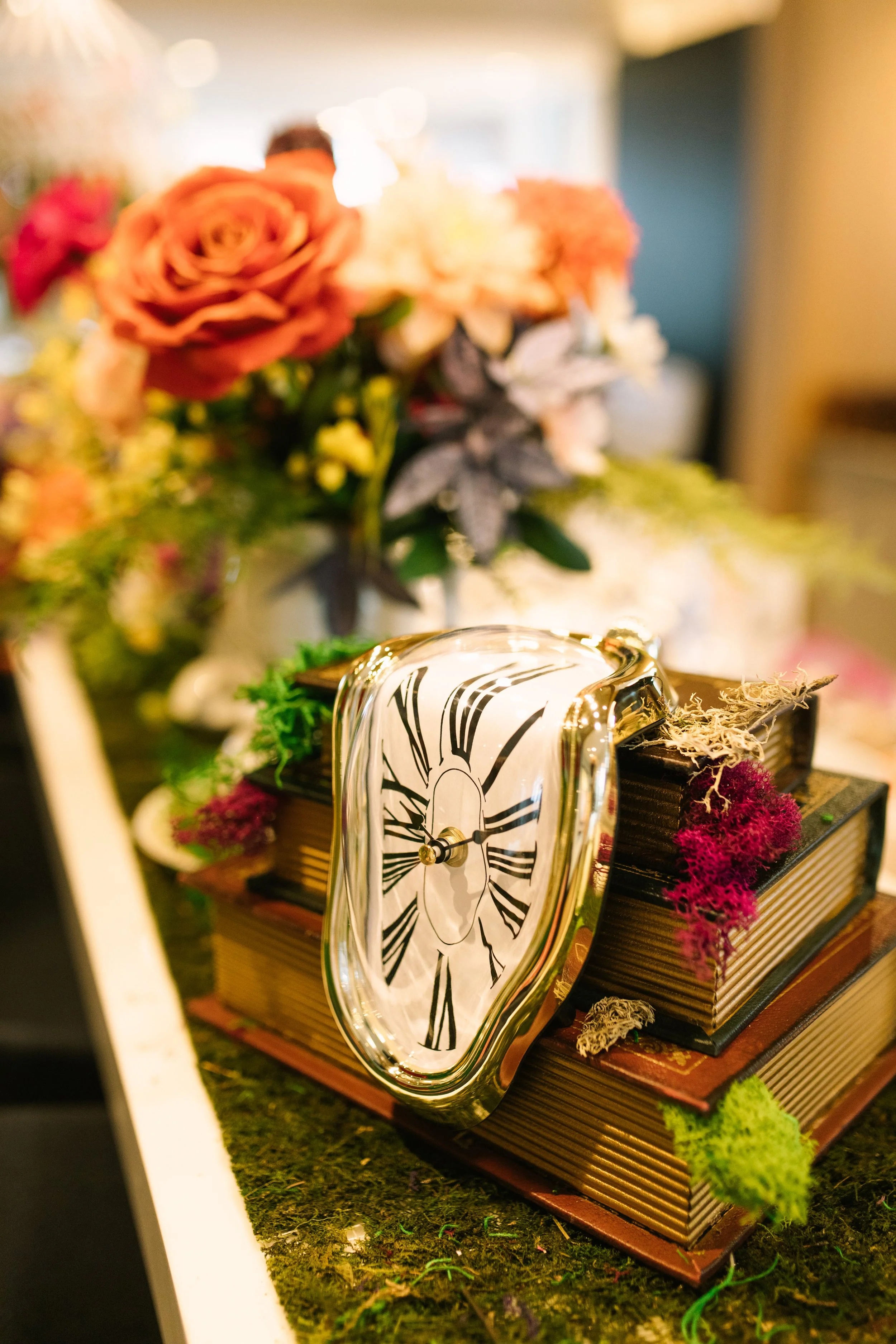 A warped, melting clock resting on a stack of books, surrounded by colorful moss and an out-of-focus bouquet of flowers in the background.