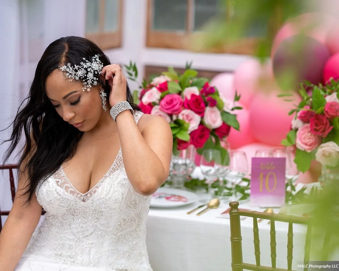 A woman in a white wedding dress with lace details, wearing a jeweled hair accessory and matching jewelry, standing at a decorated table with pink and white roses, balloons, and a table number sign.