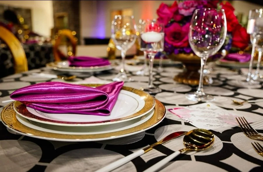 Elegant dining table set with gold-rimmed plates, a purple napkin, glasses, and a floral centerpiece featuring pink roses.