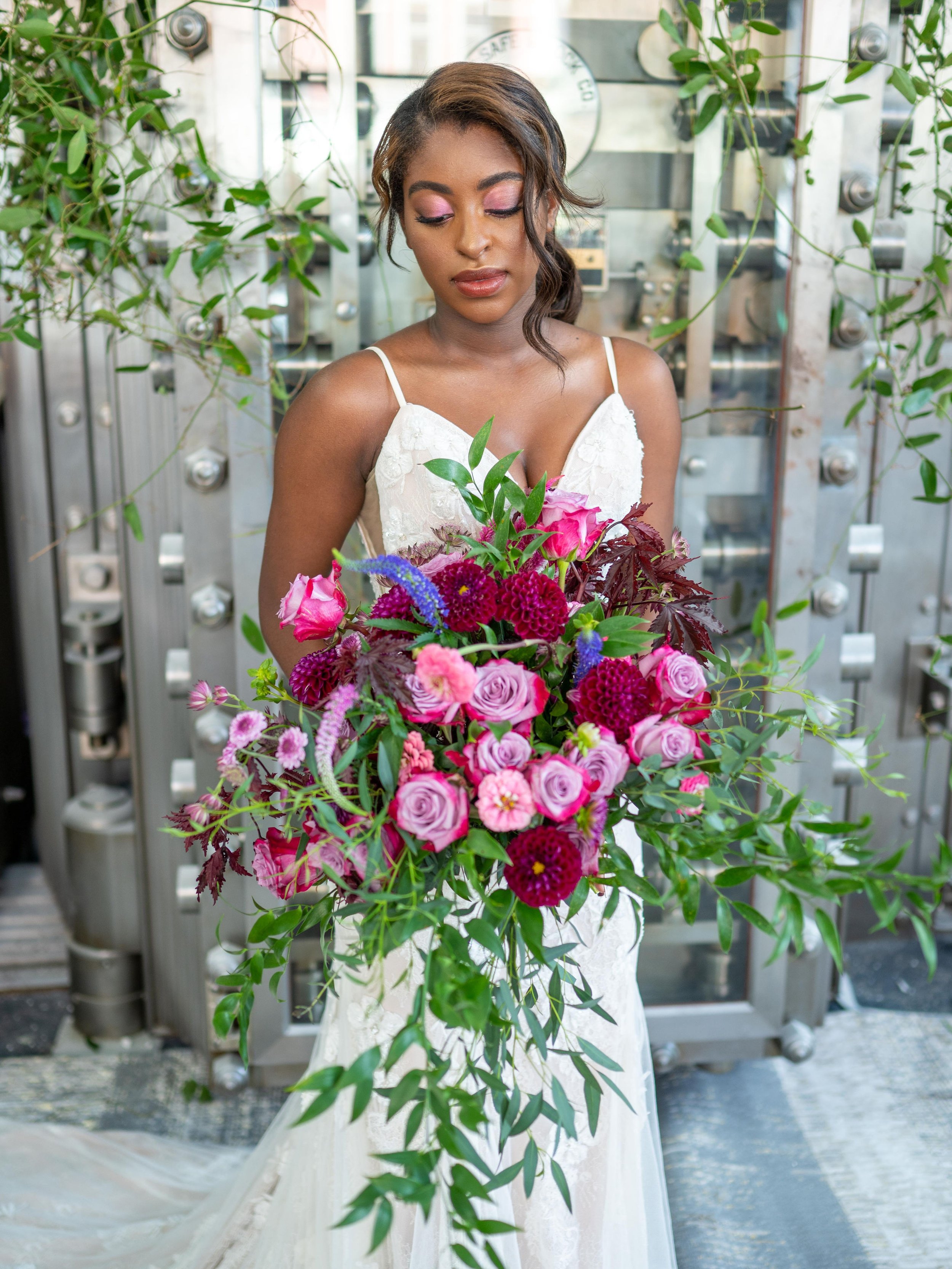 A woman in a white wedding dress holding a large bouquet of pink and purple flowers, standing in front of an industrial metal background.