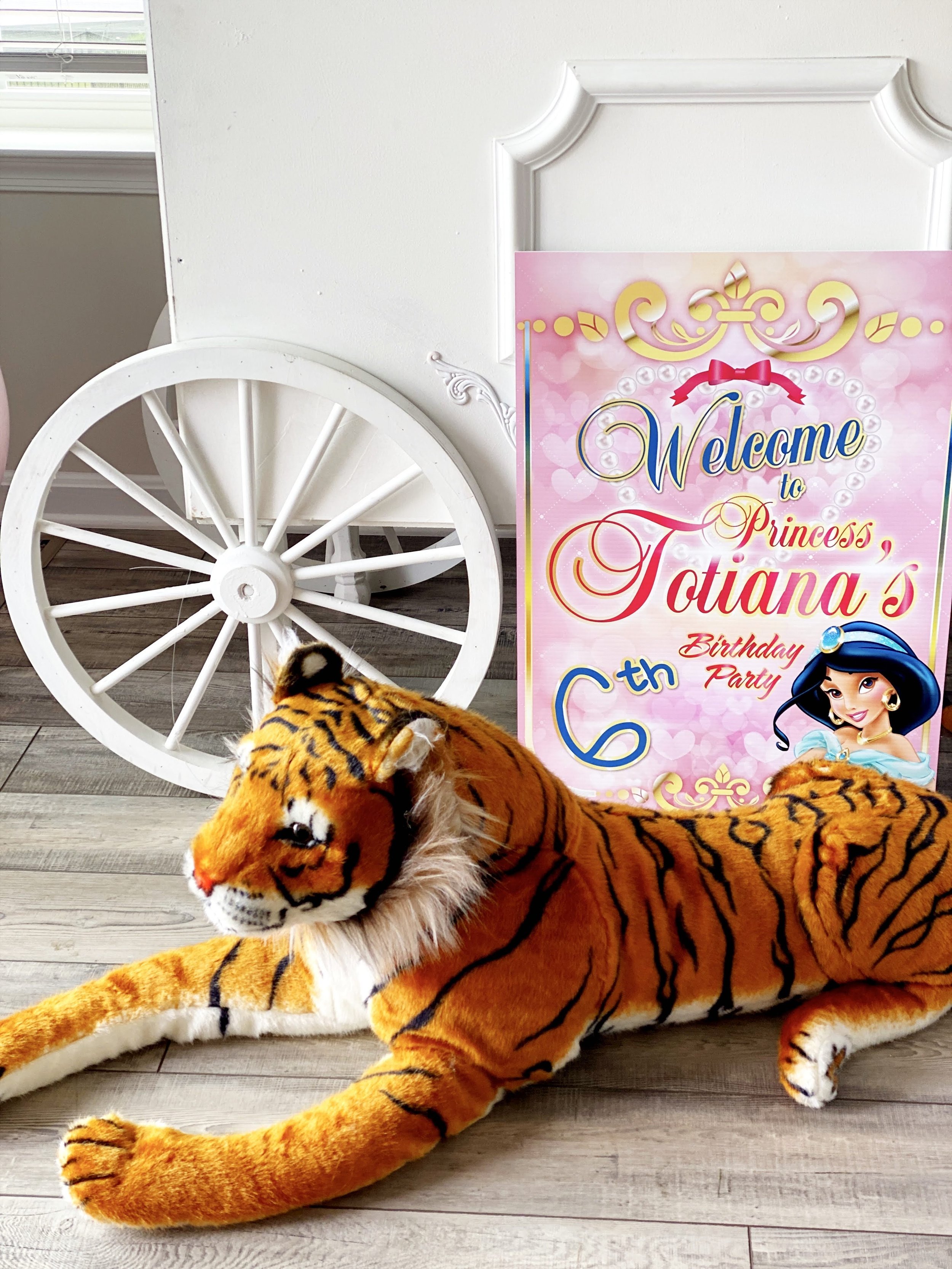 A plush tiger lying on the floor next to a pink Princess Jasmine-themed birthday sign that reads "Welcome to Princess Jotiana's 6th Birthday Party." There is a white decorative wagon wheel and part of a window in the background.