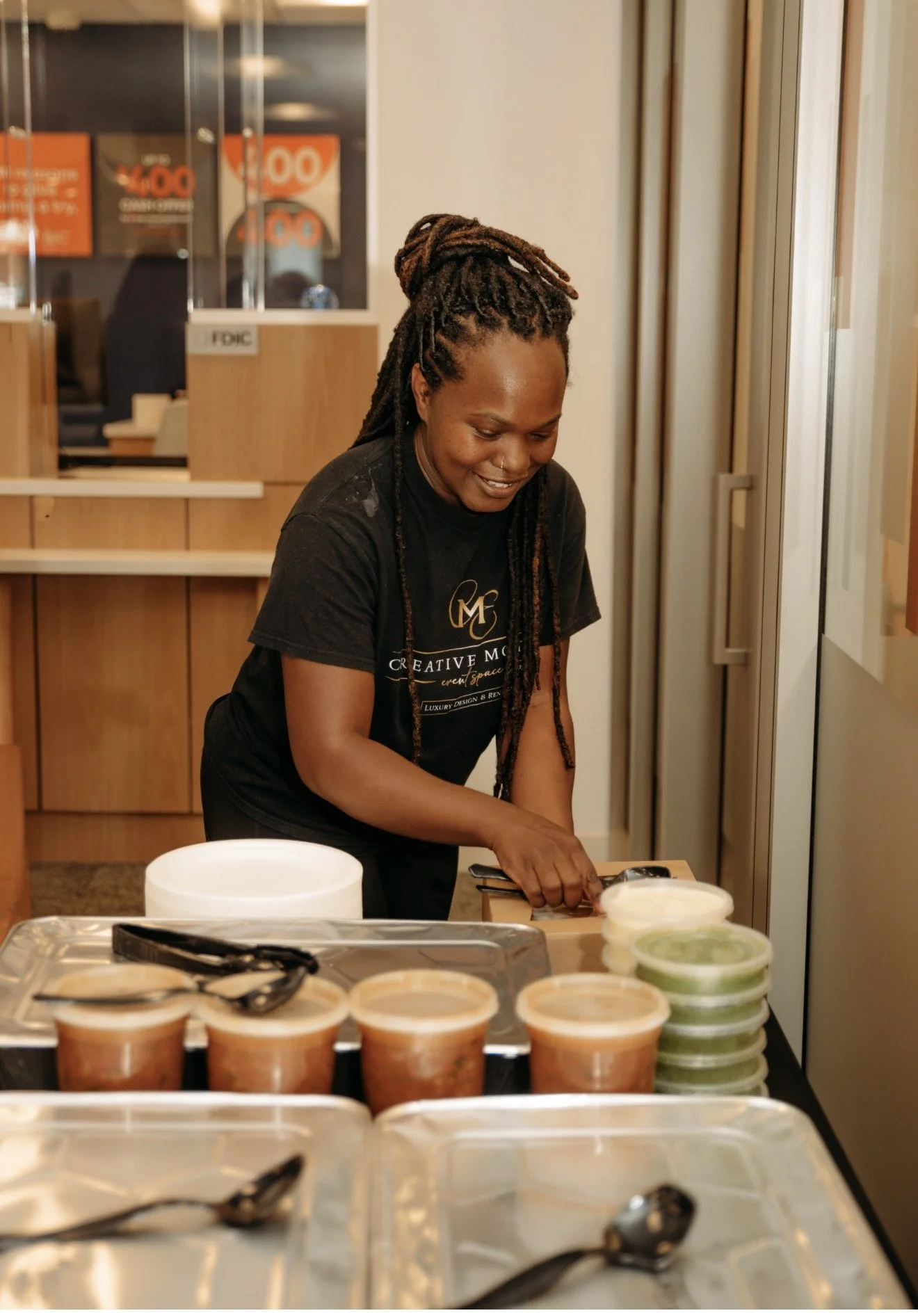 A woman with dreadlocks smiling and preparing food in a kitchen or food service area with containers and utensils on the table.