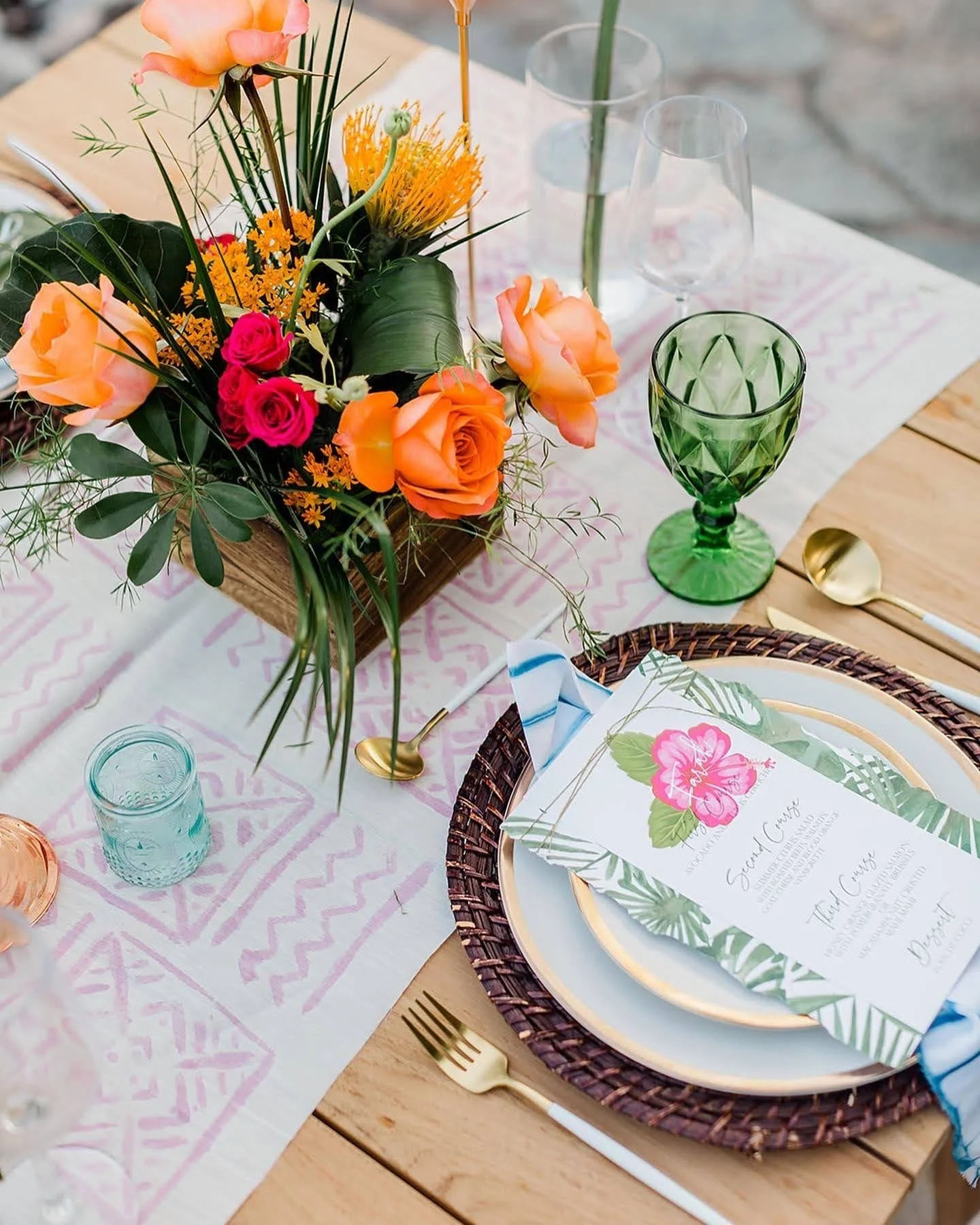 Table setting with a floral centerpiece, green and clear drink glasses, a gold spoon, gold fork, decorative plate, and a printed menu card.