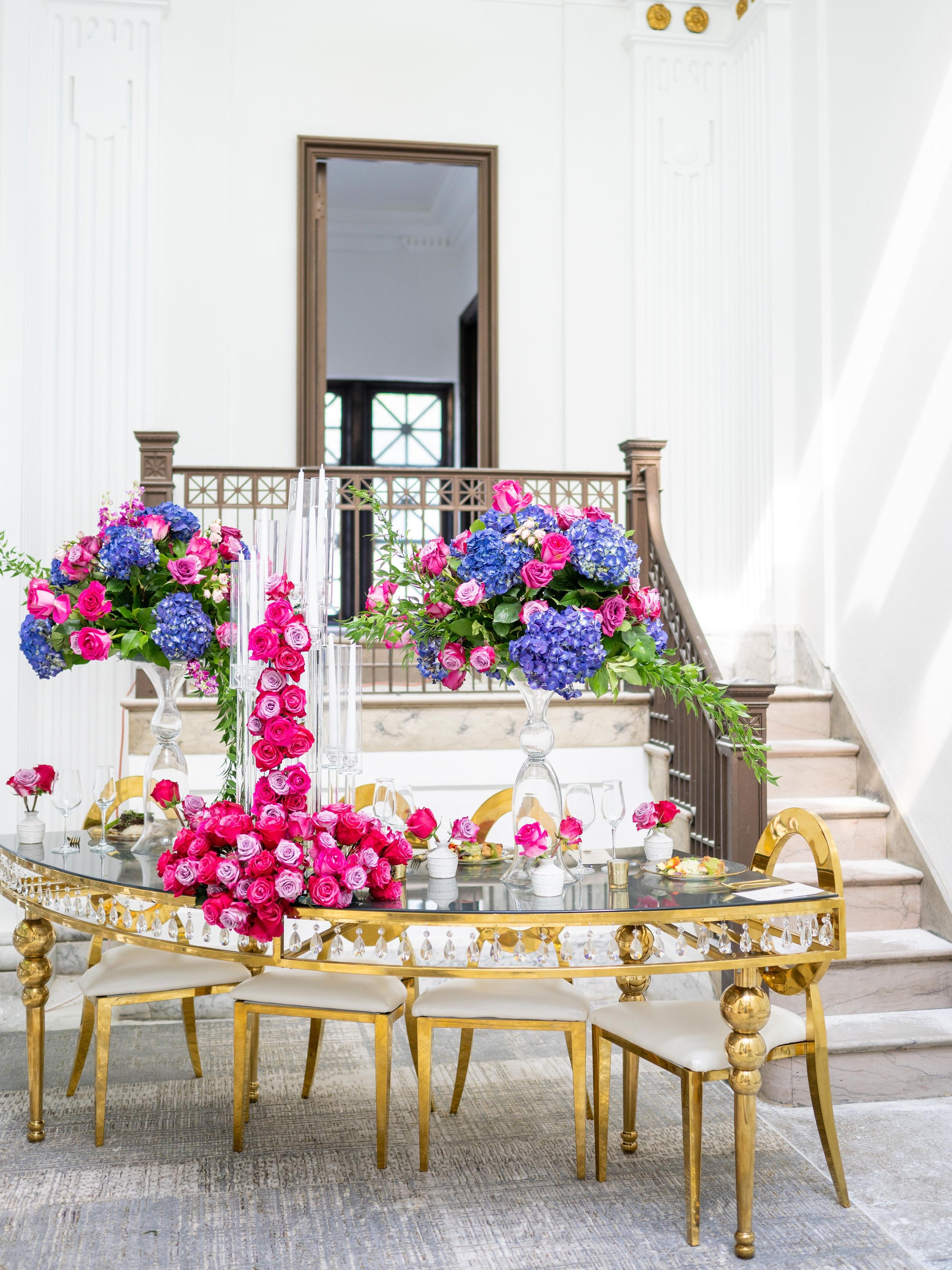 Elegant floral arrangement on a gold mirrored table with chairs in a bright, white room featuring stairs and a large mirror.