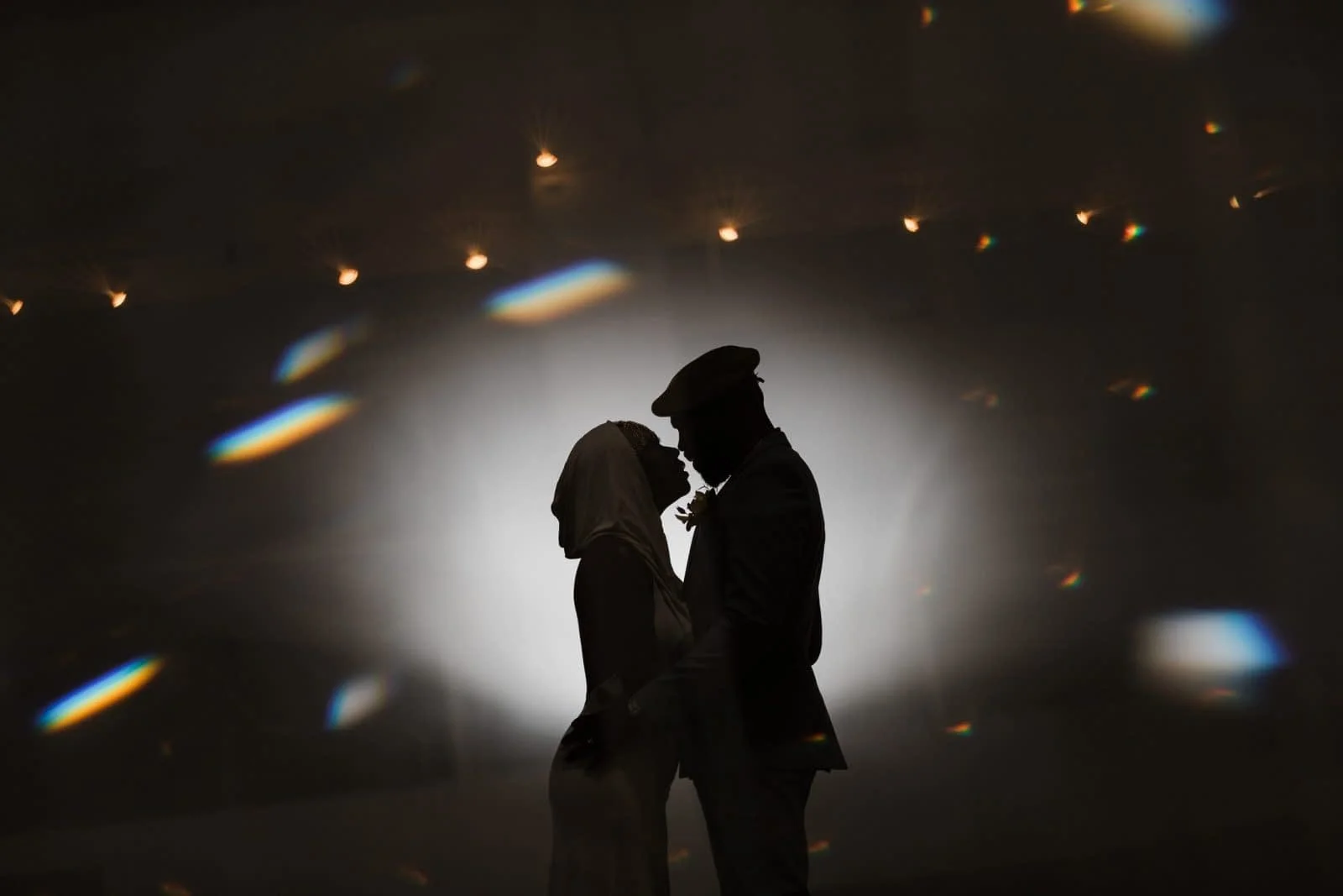 Silhouette of a couple in wedding attire, standing close and touching foreheads, with a dark background and scattered rainbow-like light reflections.