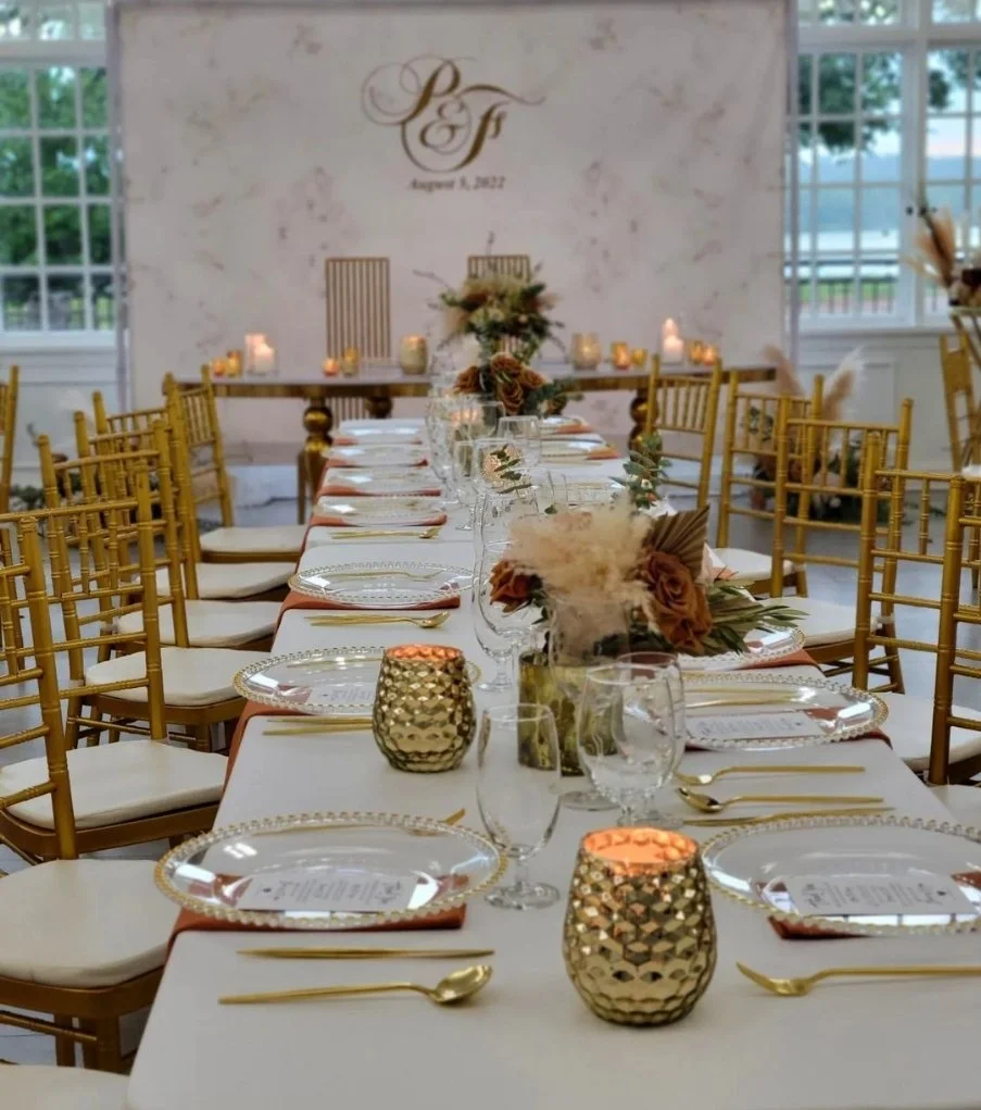 Elegant wedding reception table setup with gold chairs, floral centerpieces, candles, and gold-accented tableware, with initials and date on the wall in the background.