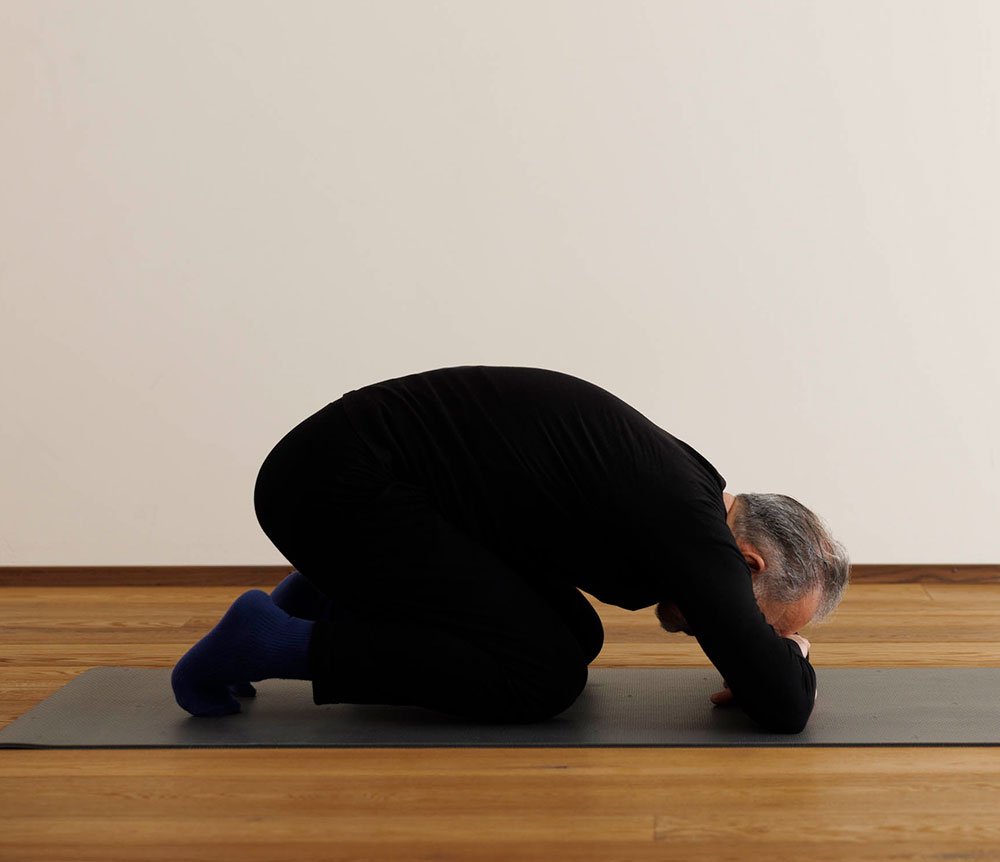 Ein älterer Mann in Schwarz Kleidung macht Yoga in einer Kniestellung auf einer Yogamatte auf einem Holzboden vor einer hellen Wand.