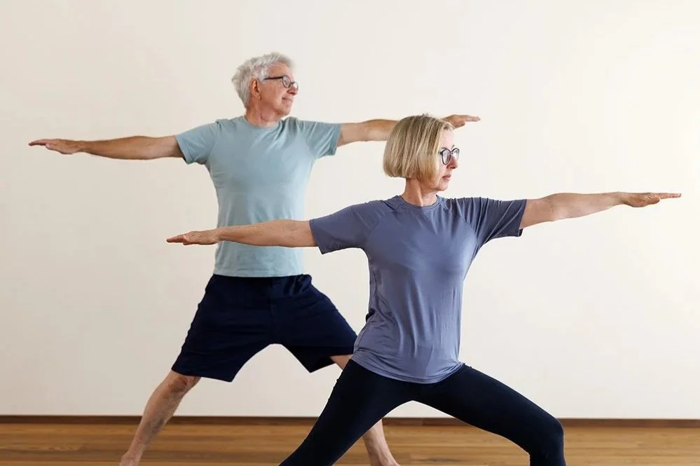 Frau in Yoga-Position auf einer Yoga-Matte, weißes T-Shirt und grüne Leggings, in einem hellen Raum mit Holz-Wand.