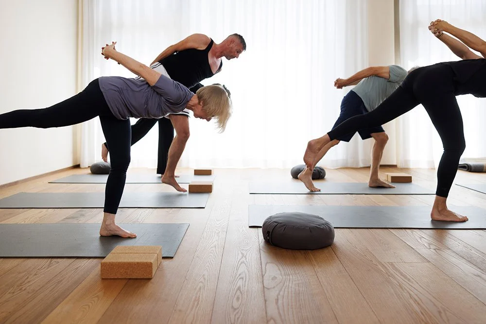 Menschen beim Yoga in einer hellen, großen Raum mit Holzboden, Mondrian-ähnlichen Mustern, großen Fenstern und Blick auf Bäume im Hintergrund.