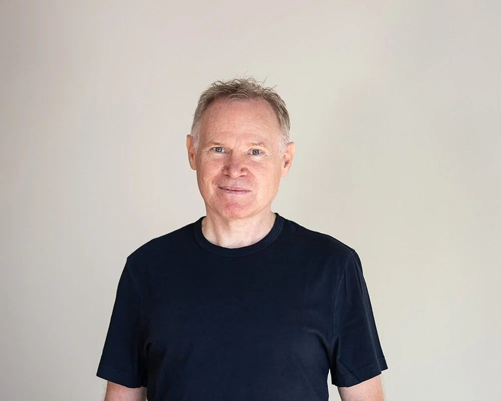 Ein Mann mit hellen Haaren, trägt ein schwarzes T-Shirt, steht vor neutralem Hintergrund, lächelt leicht.