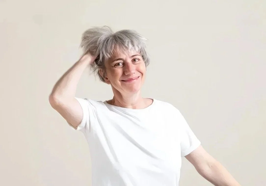 Eine Frau mit grauem, mittellangem Haar trägt ein weißes T-Shirt, lächelt und hebt eine Hand an den Kopf vor einer neutralen Wand.