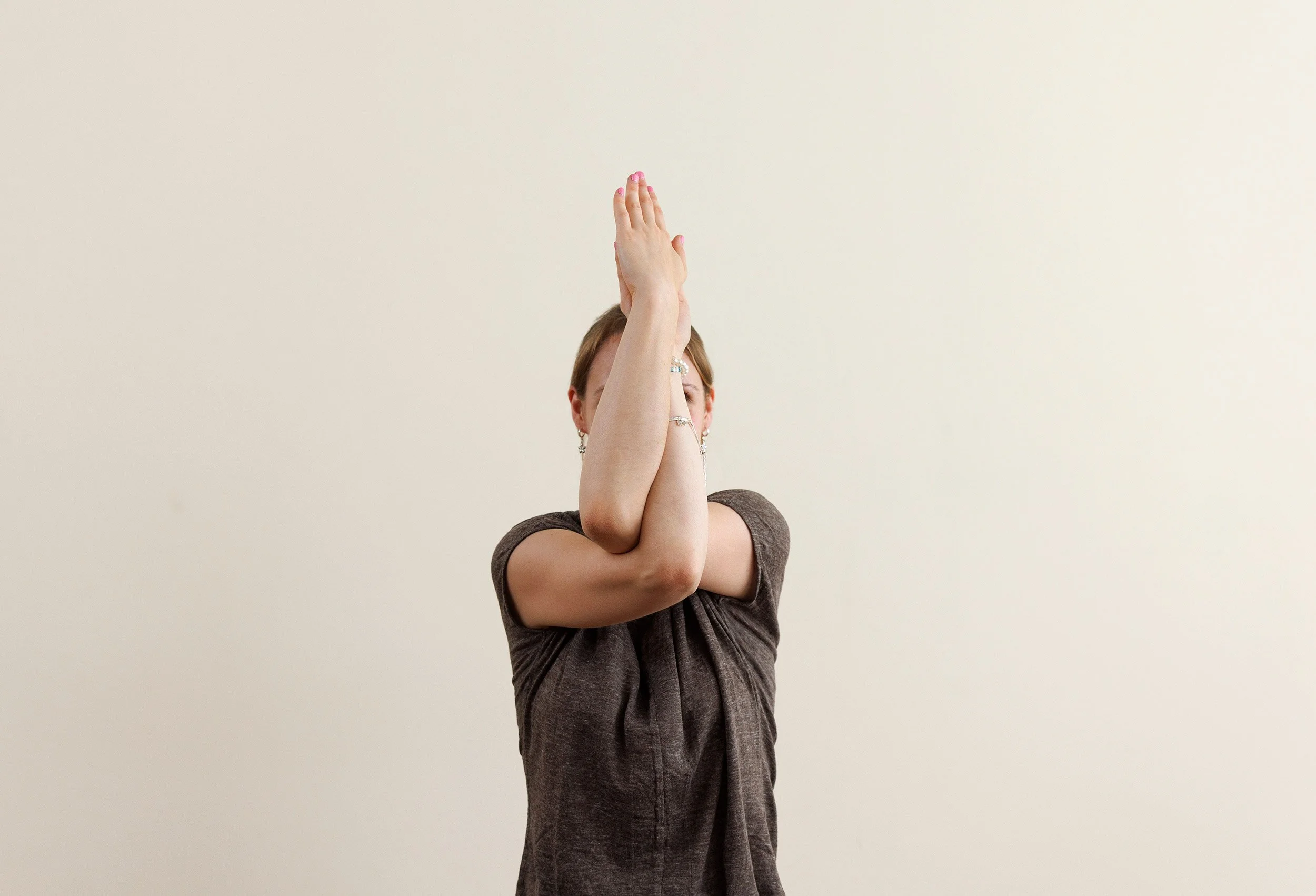 Frau, die in Yoga-Position mit verschränkten Armen und Handflächen aneinander, vor einem neutralen Hintergrund steht.