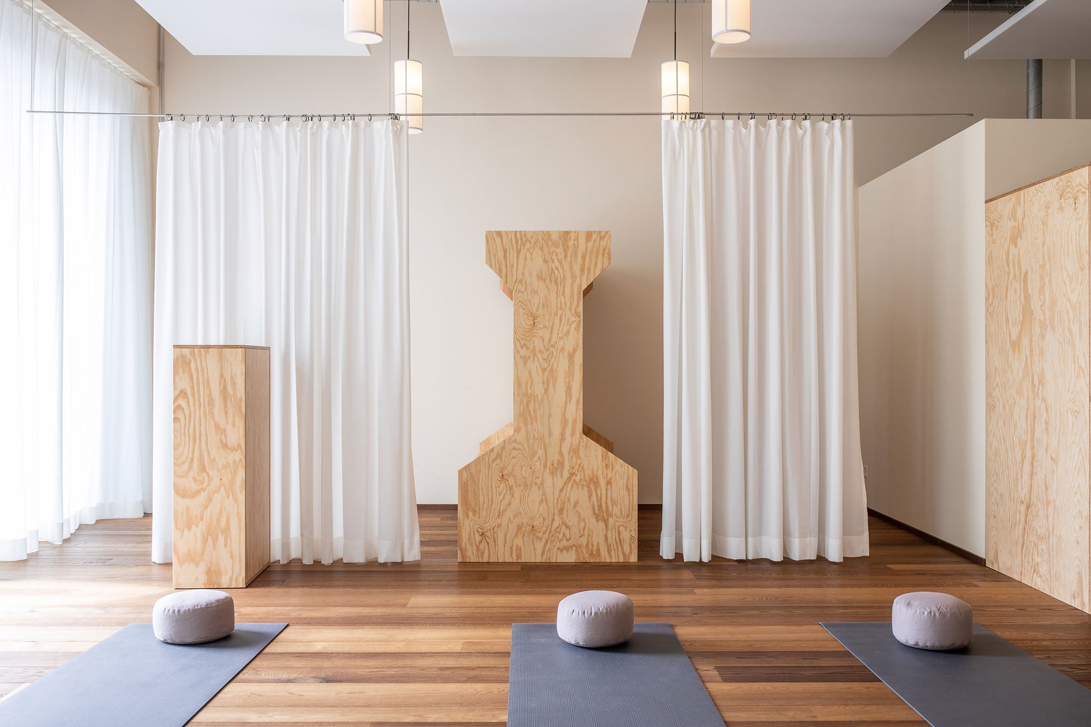 Yoga-Raum mit drei Yogamatten und Sitzkissen auf Holzboden, durch weisse Vorhänge abgeteilter Bereich, moderne Beleuchtung und helle Wände.
