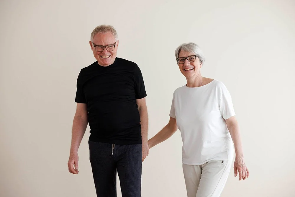 Ein älterer Mann und eine ältere Frau, beide lächelnd und haltend, stehen vor einem weißen Hintergrund.