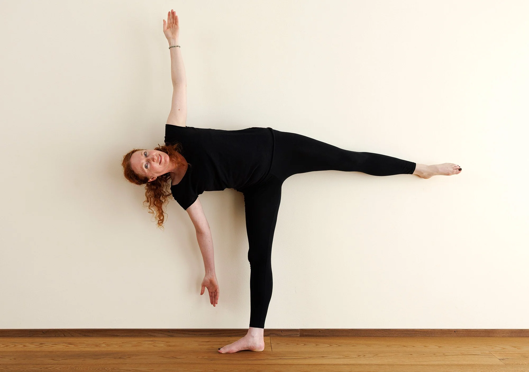 Frau in schwarzer Kleidung führt eine Seite-Krieger-Yoga-Pose aus, bei der ein Arm nach oben und der andere nach unten ausgestreckt ist, während sie auf einem weißen Boden gegen eine weiße Wand steht.