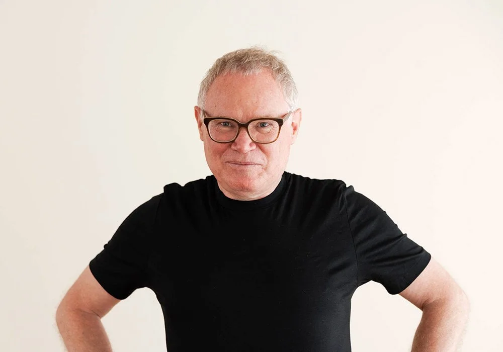 Ein älterer Mann mit Brille trägt ein schwarzes T-Shirt und steht vor einem hellen Hintergrund.