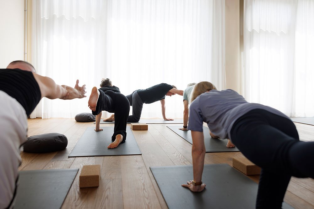 Menschen beim Yoga in einem hellen Raum mit Vorhängen vor grossen Fenstern.