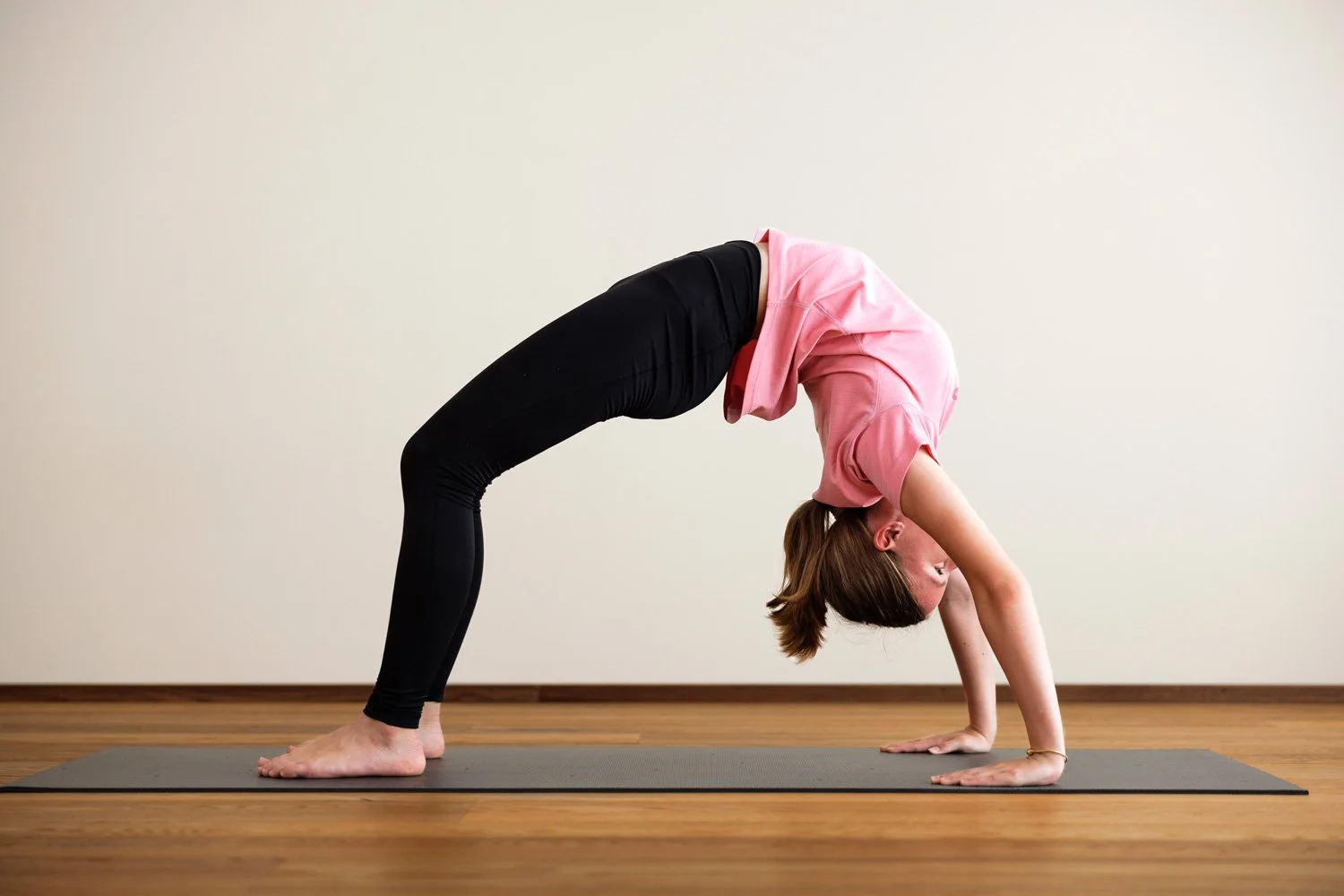 Zwei Frauen sitzen im Yoga-Raum auf Yogamatten, sprechen und lachen. Sie tragen Sportbekleidung und befinden sich bei Tageslicht in einem hellen Raum mit großen Fenstern und einer Topfpflanze.