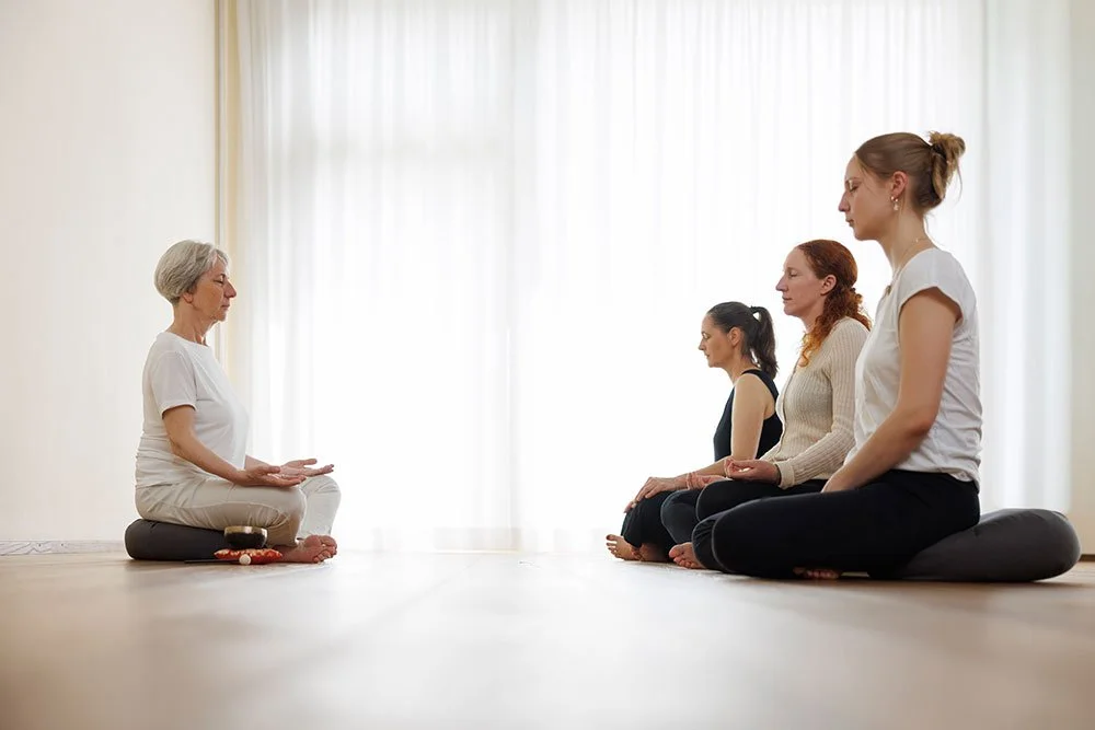 Vier Frauen sitzen im Schneidersitz in einer Meditationshaltung gegenüber einer älteren Frau, die vor ihnen ebenfalls im Lotussitz Meditationshaltung einnimmt, in einem hell erleuchteten Raum.
