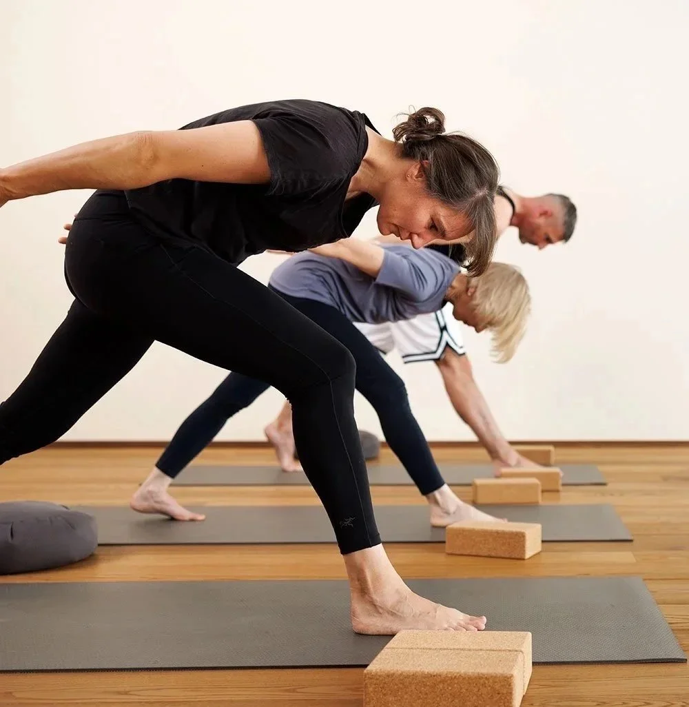 Menschen beim Yoga in einer hellen, großen Raum mit Holzboden, Mondrian-ähnlichen Mustern, großen Fenstern und Blick auf Bäume im Hintergrund.